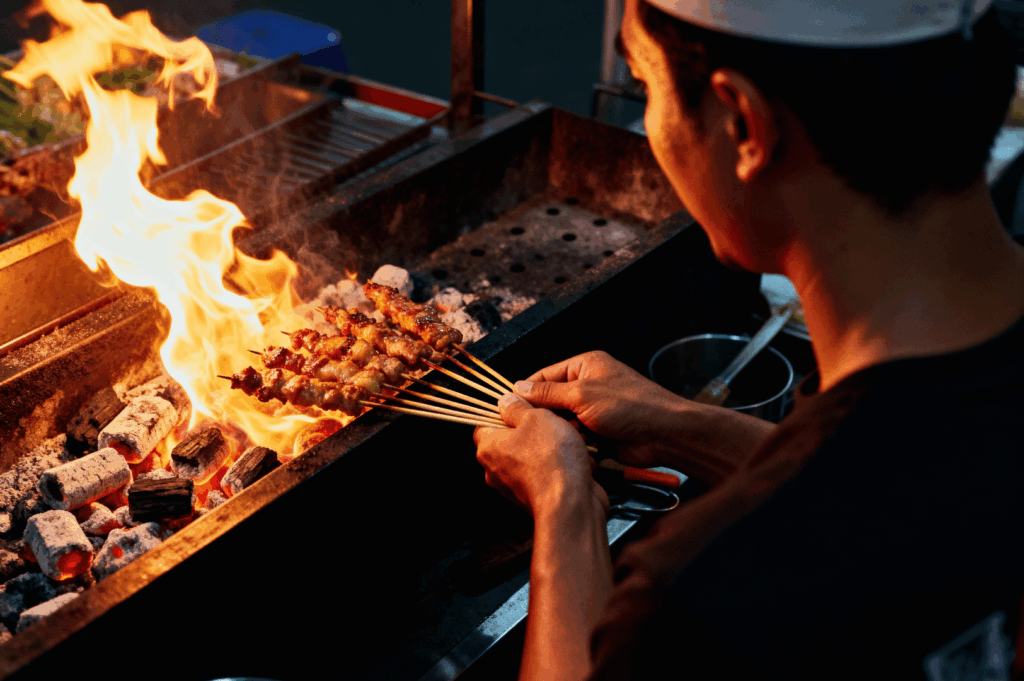 A satay master in a white head covering steadies multiple skewers over a roaring charcoal grill, flames flickering beneath the metal grate. The meat sizzles as smoke rises into the humid night, framing the chef’s focused posture and deliberate movements. It’s a moment of tactile precision and ancestral rhythm—where fire, patience, and trust converge in the art of street-side grilling.