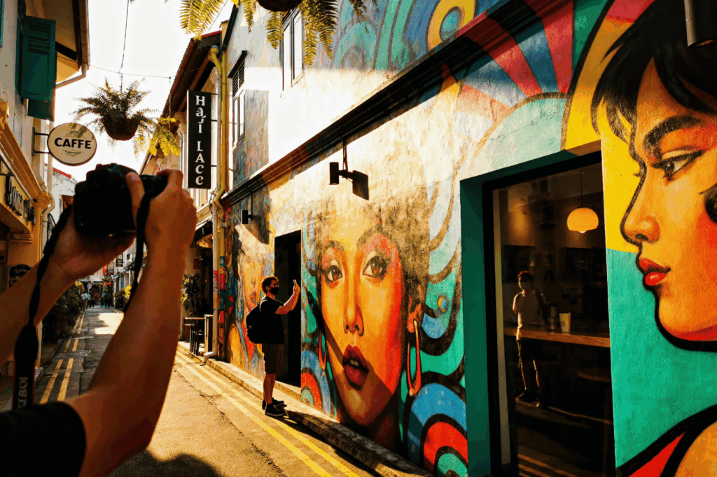 Warm light bathes Haji Lane’s mural-lined alley as a photographer captures a subject posing before expressive street art—blue-haired woman with red lips and bold makeup. Narrow street framed by café signage and layered textures, ideal for showcasing contrast, character, and the intimacy of early morning or golden hour shoots.