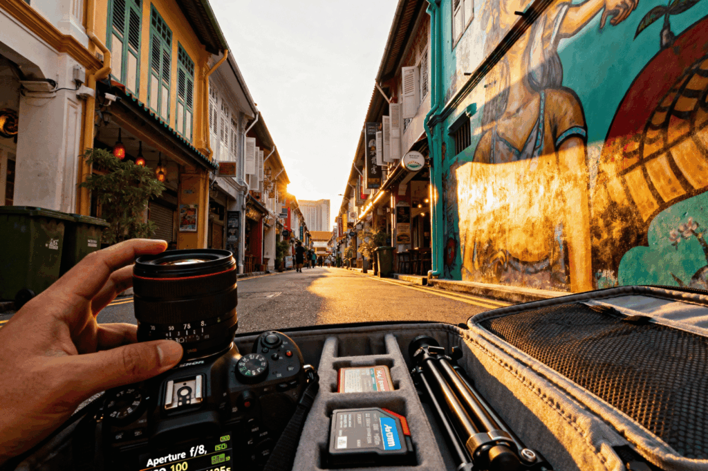 Sunset over Haji Lane, Singapore—camera gear laid out in foreground, framing a colonial-style street bathed in golden light. Photographer’s hand holds a lens, with camera display showing f/1.8 and ISO 100. Murals, lanterns, and pastel buildings line the alley, offering ideal conditions for low-light street photography and gear-focused compositions.