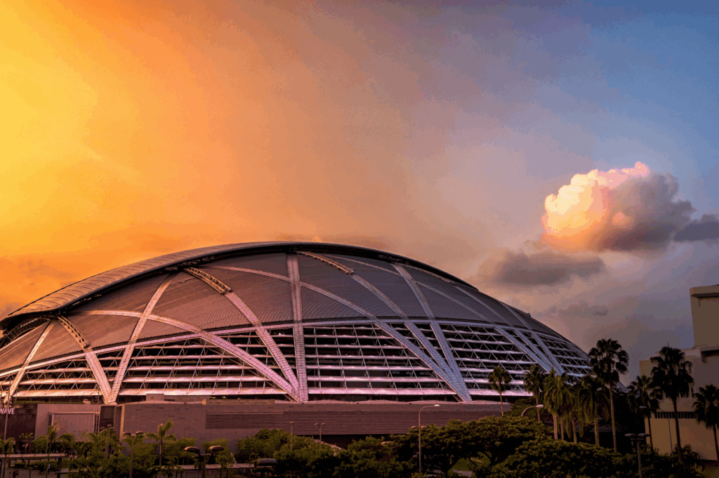 Dome-shaped stadium in Singapore framed by palm trees and dramatic sunset sky; geometric metal-and-glass architecture contrasts with vivid orange and deep blue hues above, as illuminated clouds enhance the grandeur of the structure—capturing the interplay of modern design and golden hour light.