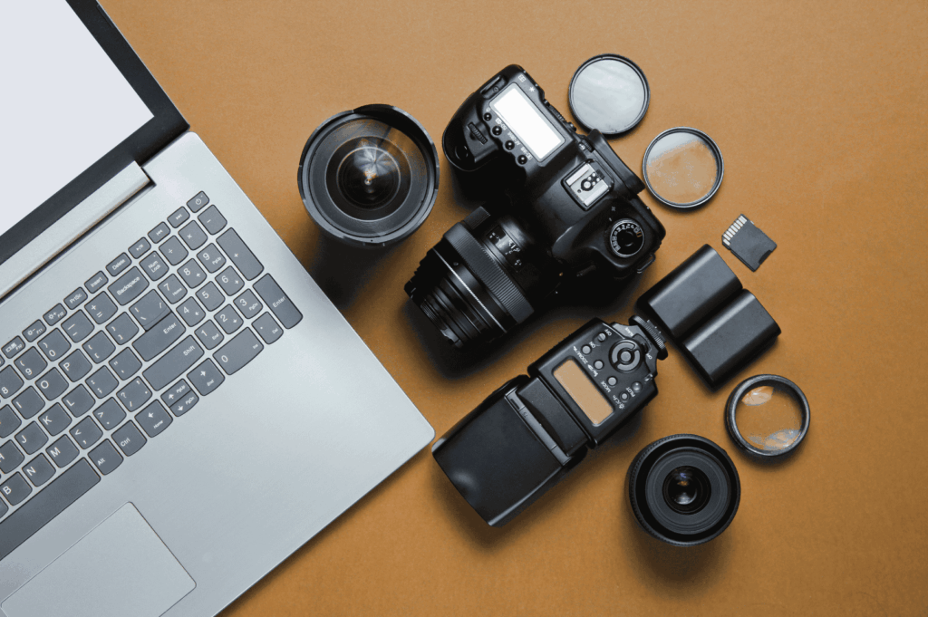 Flat lay of essential photography gear on a warm-toned surface, including a DSLR camera, lenses, filters, flash unit, memory card, and silver laptop—evoking the meticulous preparation behind capturing Singapore’s skyline at sunset during golden hour.
