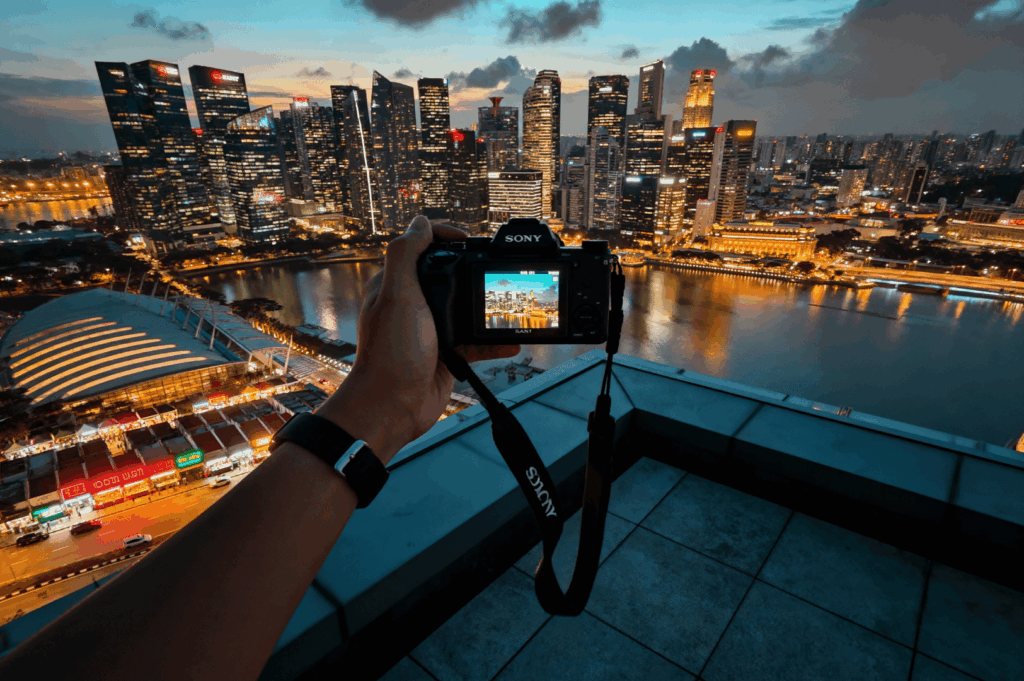 Close-up of a hand holding a Sony Alpha camera, capturing Singapore’s skyline at dusk with glowing city lights and a moody sky
