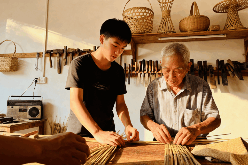 Elder and younger craftspeople shaping bamboo strips into a basket inside a cluttered workshop, illustrating the intergenerational skills of Singapore’s traditional trades.