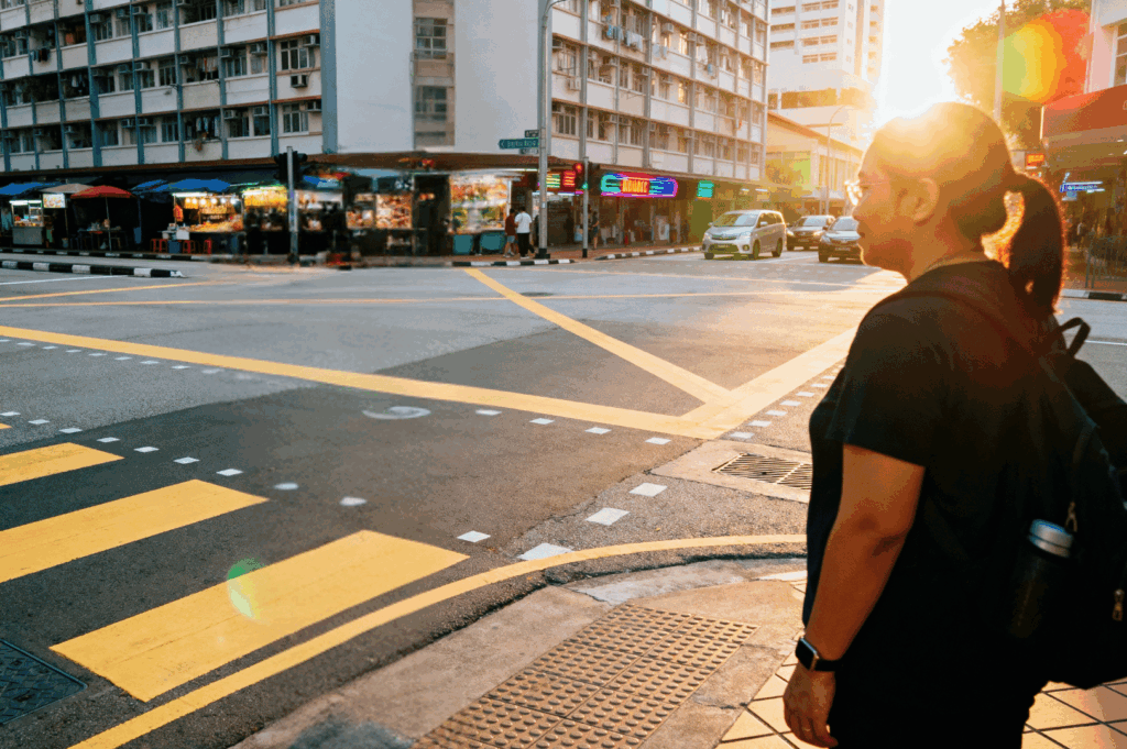 Singaporean pedestrian waiting at sunset-lit intersection with tactile paving and food stalls in view—capturing ethical street photography in everyday urban life.