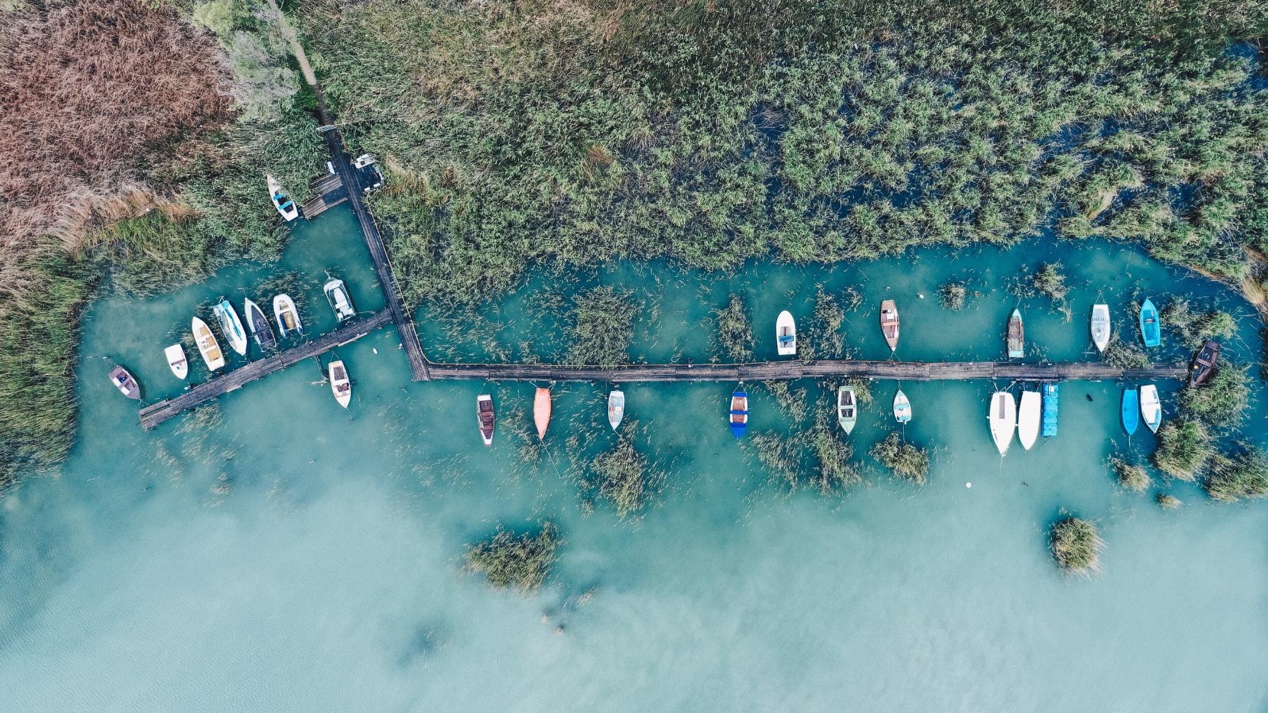  This is an overhead drone shot capturing a small, rustic wooden dock extending from a shoreline dense with tall reeds and marsh vegetation. Numerous small boats are moored along the structure in the striking, clear turquoise-blue water of a lake or bay.