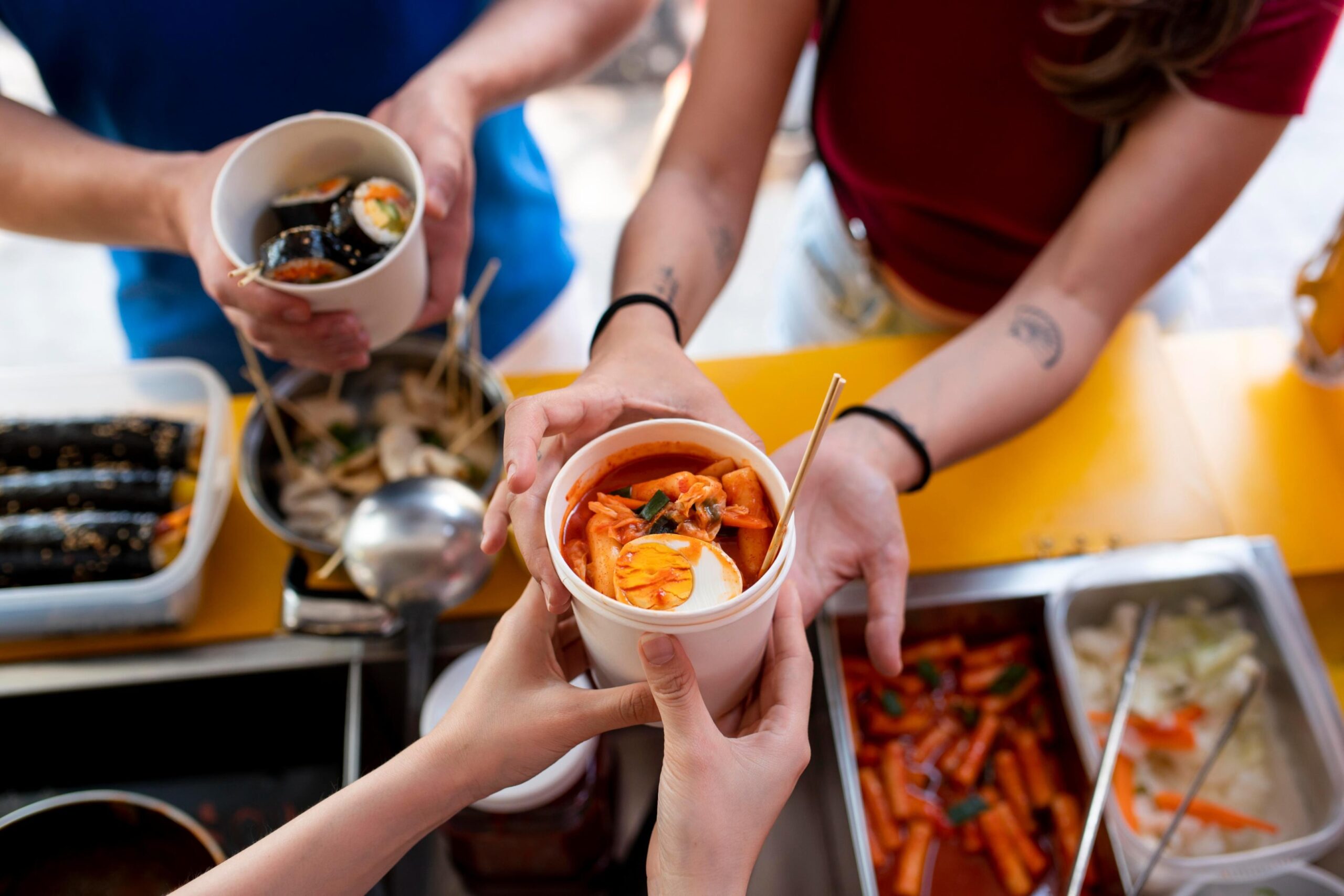  This image shows a close-up, bustling view of a transaction at a street food stall, as a customer receives a white paper cup filled with a spicy-looking stew or sauce containing a hard-boiled egg half, possibly Tteokbokki.