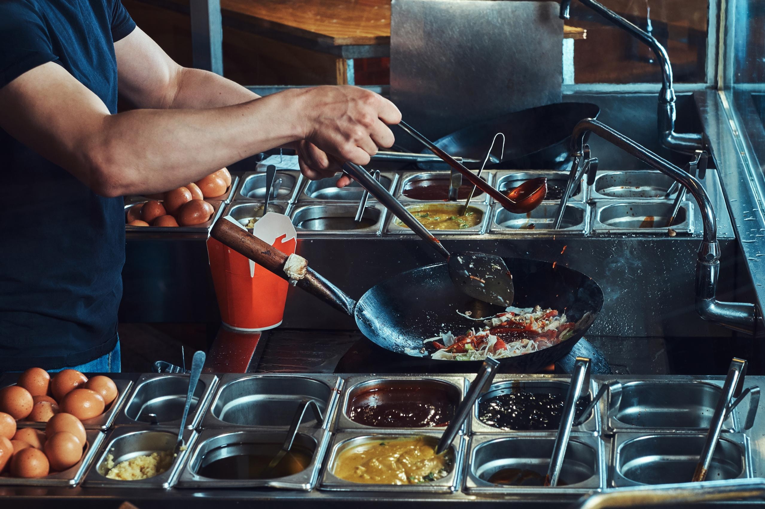  This image shows a chef preparing a dish in a large wok over high heat in a commercial kitchen setting, with their hands visible adding ingredients from a ladle. 