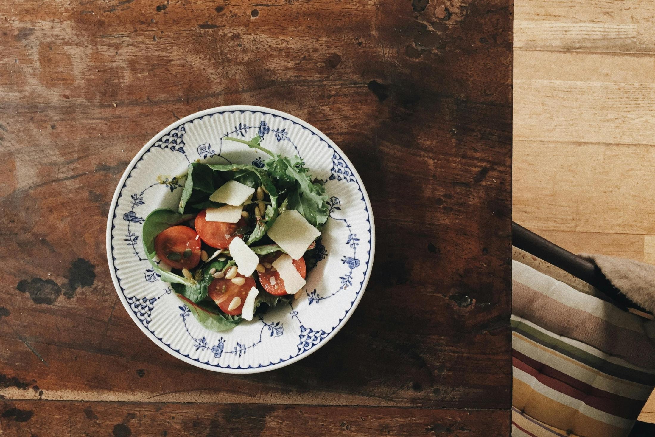 A vibrant salad of leafy greens, halved cherry tomatoes, and shaved cheese is served in a blue and white patterned bowl. The dish sits on a weathered wooden table, capturing a rustic and fresh aesthetic from an overhead perspective.