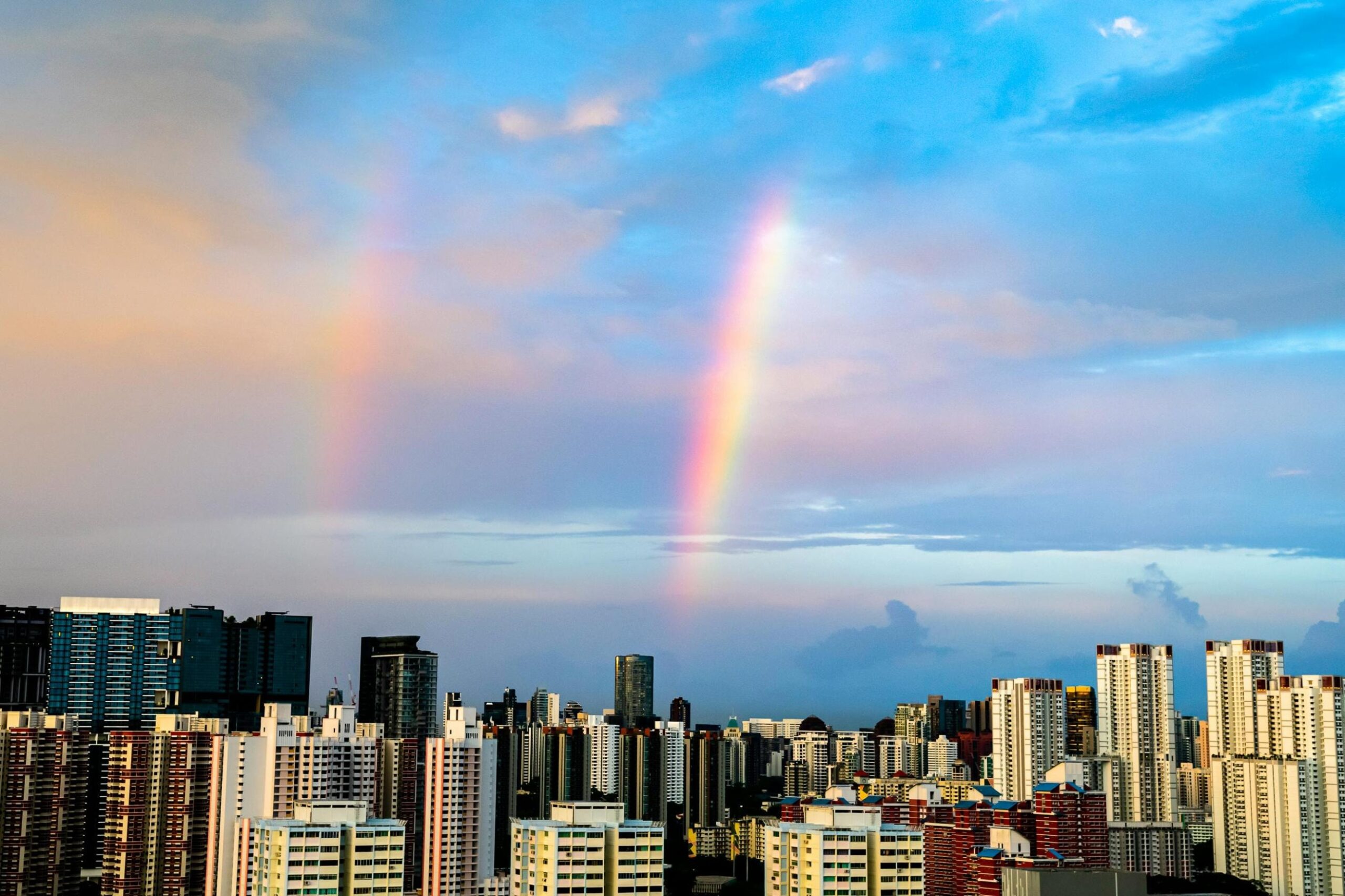 This image captures two vertical segments of a rainbow piercing through soft clouds against a blue and pink-hued evening sky. Beneath this colorful atmospheric display, a dense panoramic skyline of white and terracotta-colored high-rise apartment buildings stretches across the horizon.