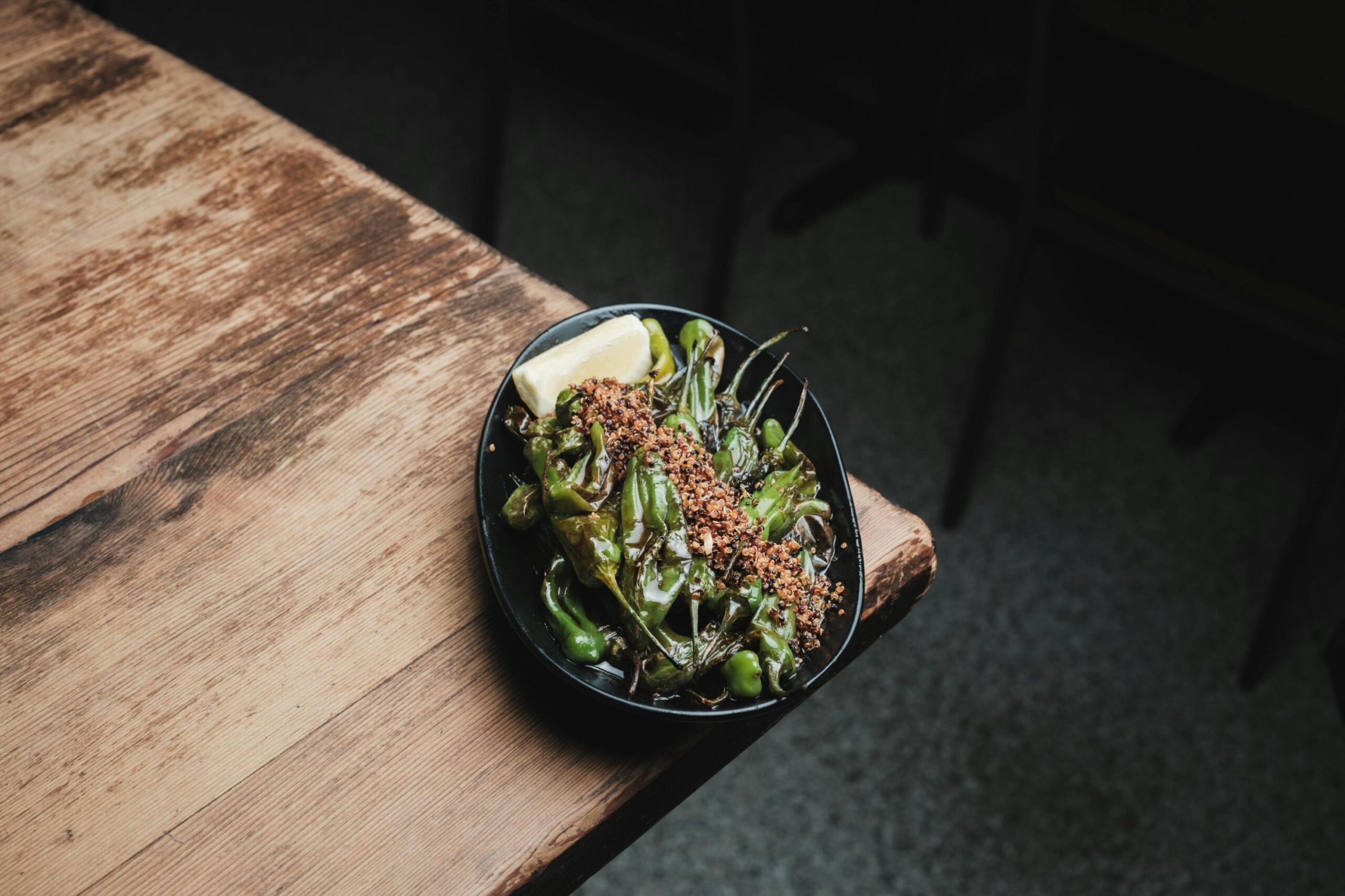 A small black plate sits on the edge of a rustic wooden table, holding a pile of blistered green peppers topped with a line of toasted crumbs and a lemon wedge.