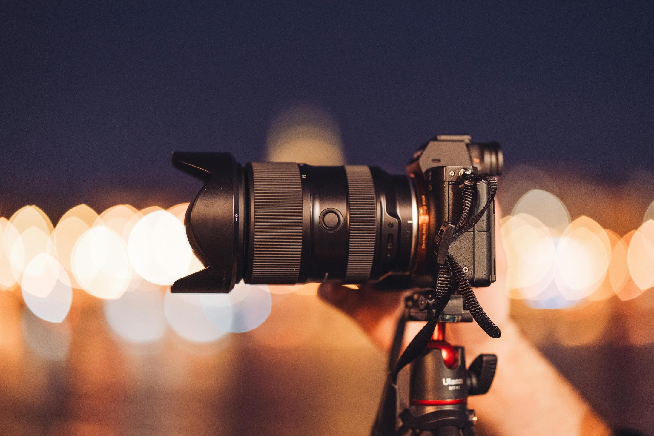 This photo depicts a black digital camera equipped with a long zoom lens and lens hood, mounted on a tripod while being steadied by a hand. The scene is set against a dark blue twilight background where distant city lights are rendered as large, warm bokeh circles.