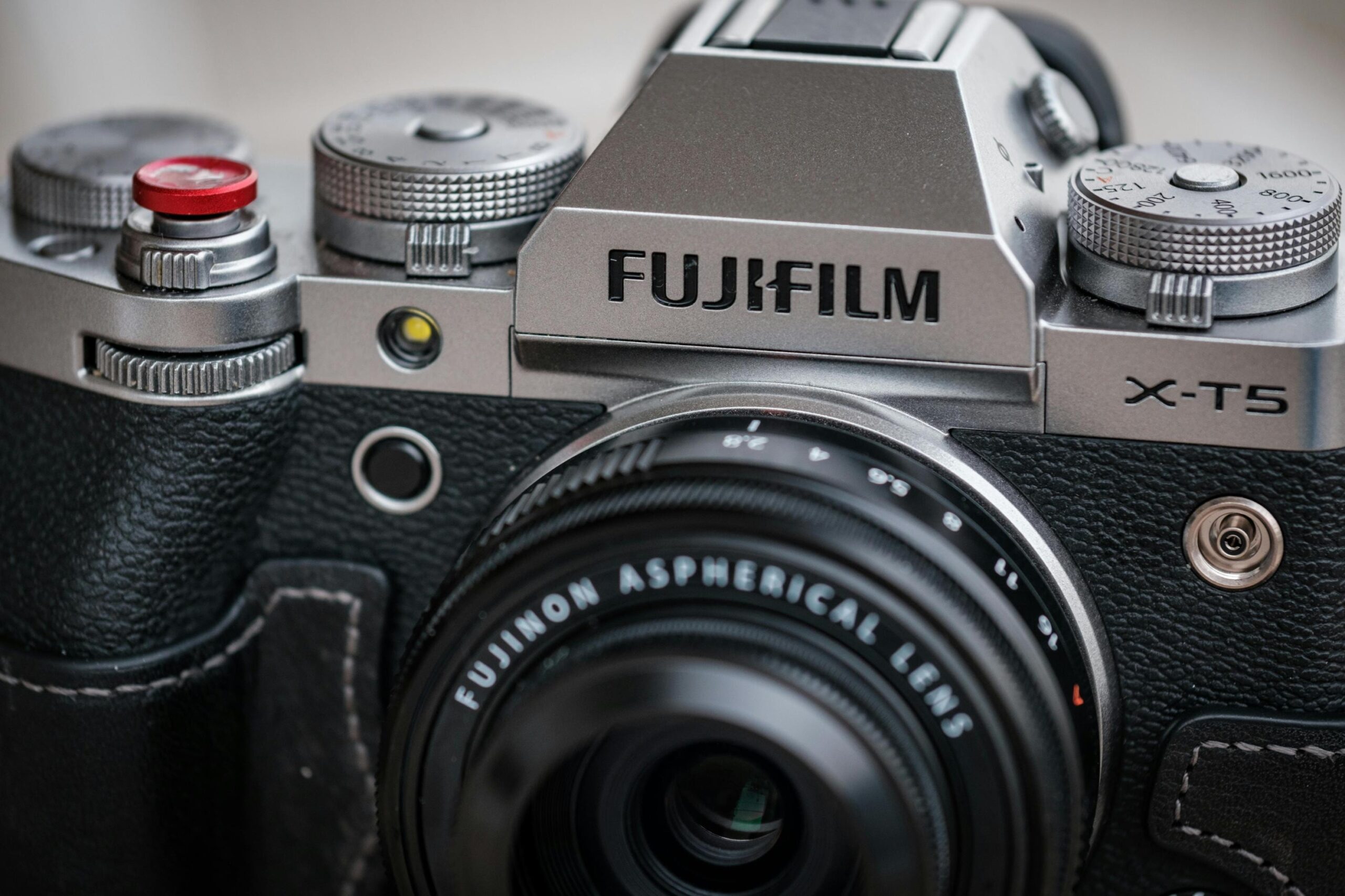 This close-up photograph captures the details of a silver Fujifilm X-T5 camera, highlighting its retro design with textured black grip material and analog-style manual dials on the top plate. The image features a Fujinon Aspherical lens attached to the body, along with a distinctive red soft-release button on the shutter.