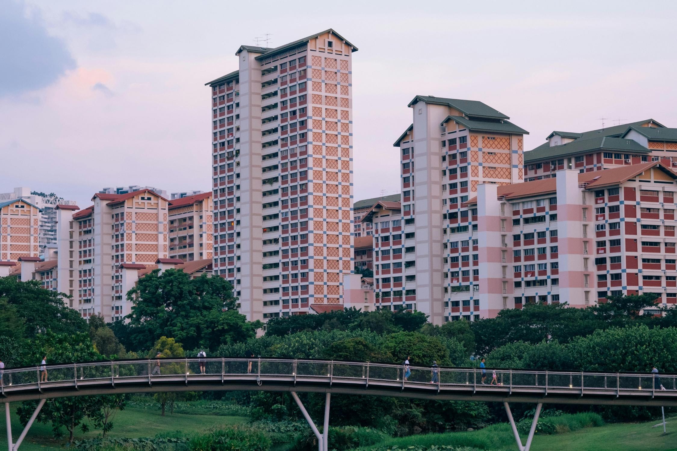 A cluster of high-rise apartment blocks characterized by beige walls, red geometric accents, and green pitched roofs dominates the background. In the foreground, a curved pedestrian bridge spans a lush park, with several people walking along the path amidst the greenery.