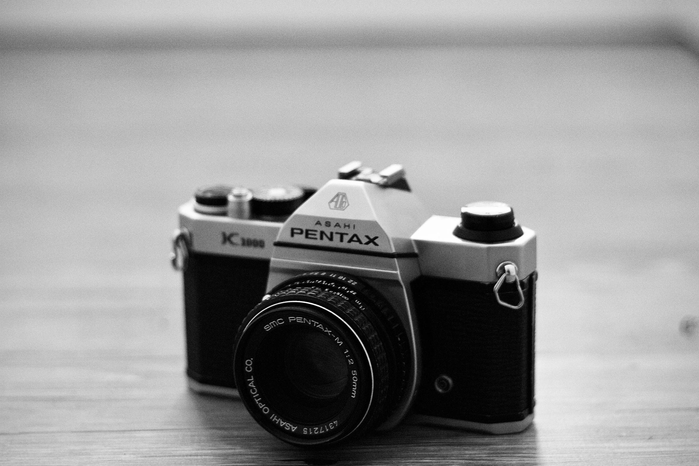 This black and white close-up features a vintage Asahi Pentax K1000 camera resting on a textured wooden surface. The shallow depth of field emphasizes the sharp details of the 50mm lens and mechanical dials against a softly blurred background, highlighting the classic analog design.