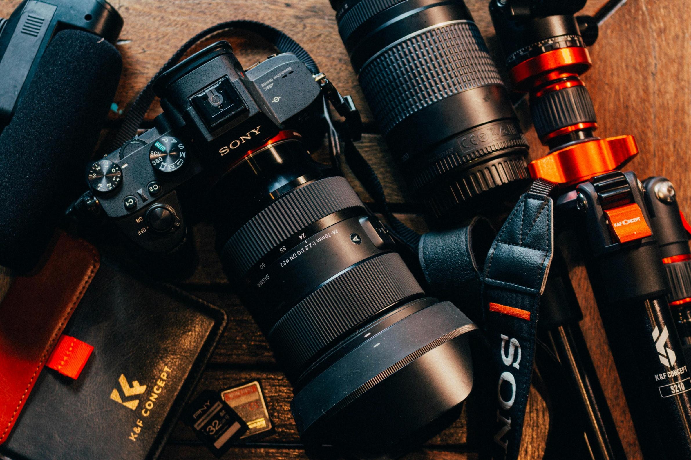  A collection of professional photography equipment, including a Sony mirrorless camera with a Sigma lens, is spread out across a dark wooden surface. The gear is accompanied by accessories like a tripod, a leather wallet, and memory cards, all captured in a warm, moody overhead shot.