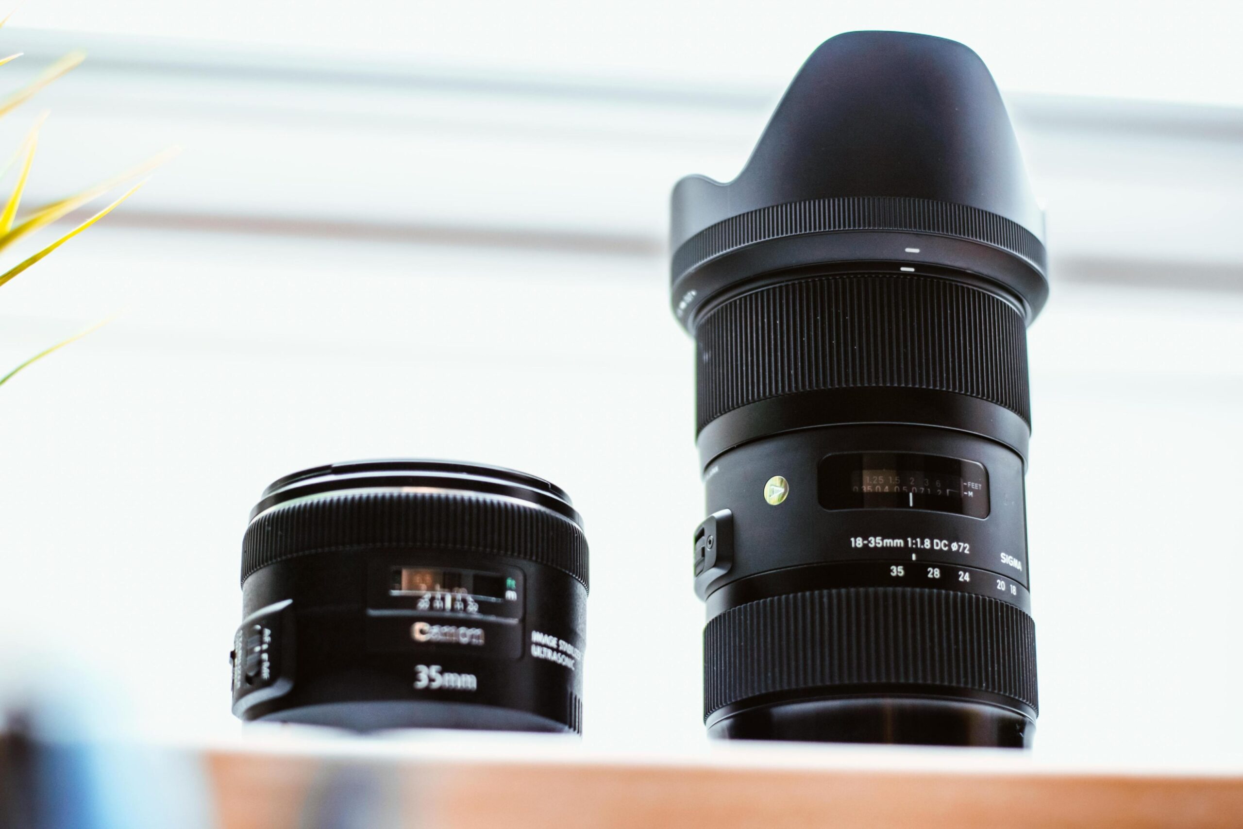  Two camera lenses, a compact Canon 35mm and a larger Sigma 18-35mm with a lens hood, stand side-by-side on a light-colored surface. The shot is taken from a low angle against a bright, softly blurred background, emphasizing the different sizes and designs of the optics.