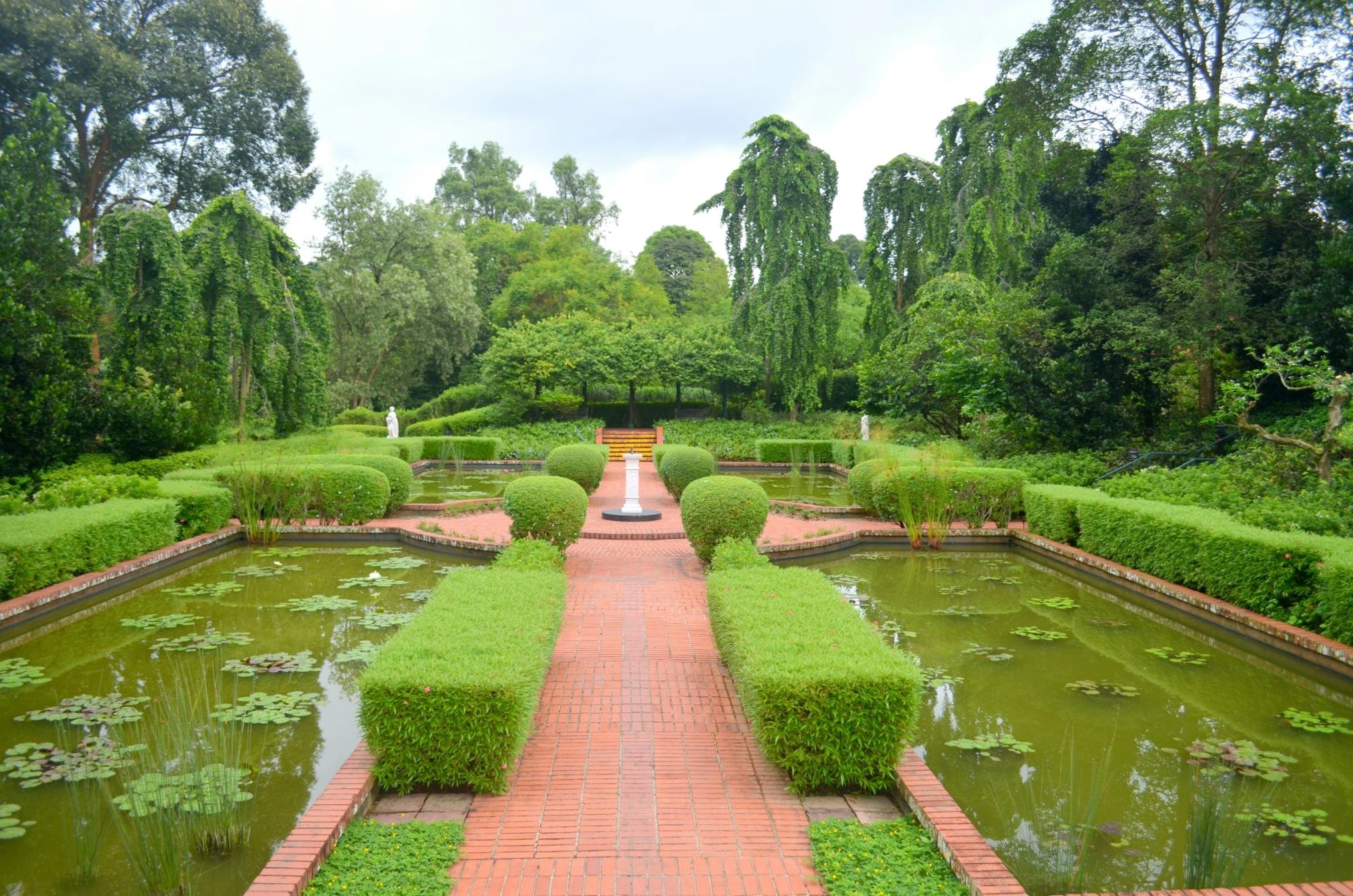 A symmetrical red brick pathway cuts through a formal garden, flanked by rectangular lily ponds and neatly trimmed green hedges. The walkway leads the eye toward a central white pedestal and stairs, surrounded by a backdrop of lush, towering trees.