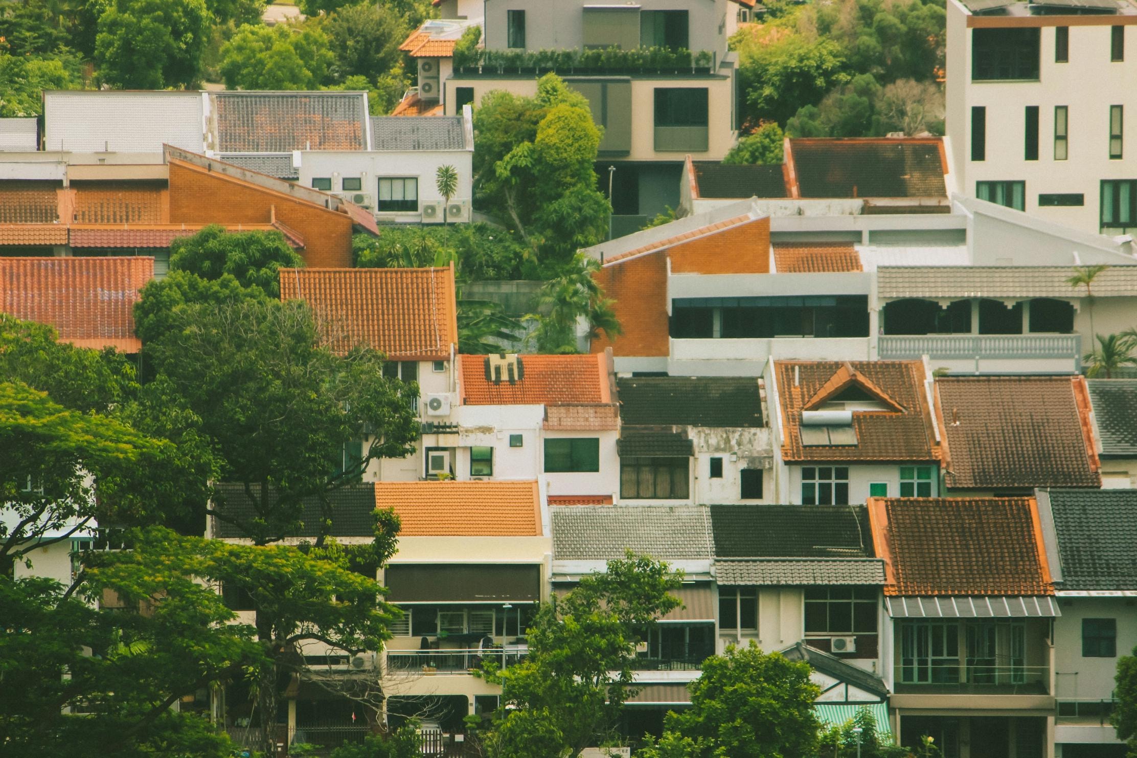 A dense residential neighborhood features a patchwork of terraced houses with traditional orange tiled roofs alongside modern white geometric structures. Lush green trees are interspersed heavily throughout the scene, softening the appearance of the tightly packed buildings.