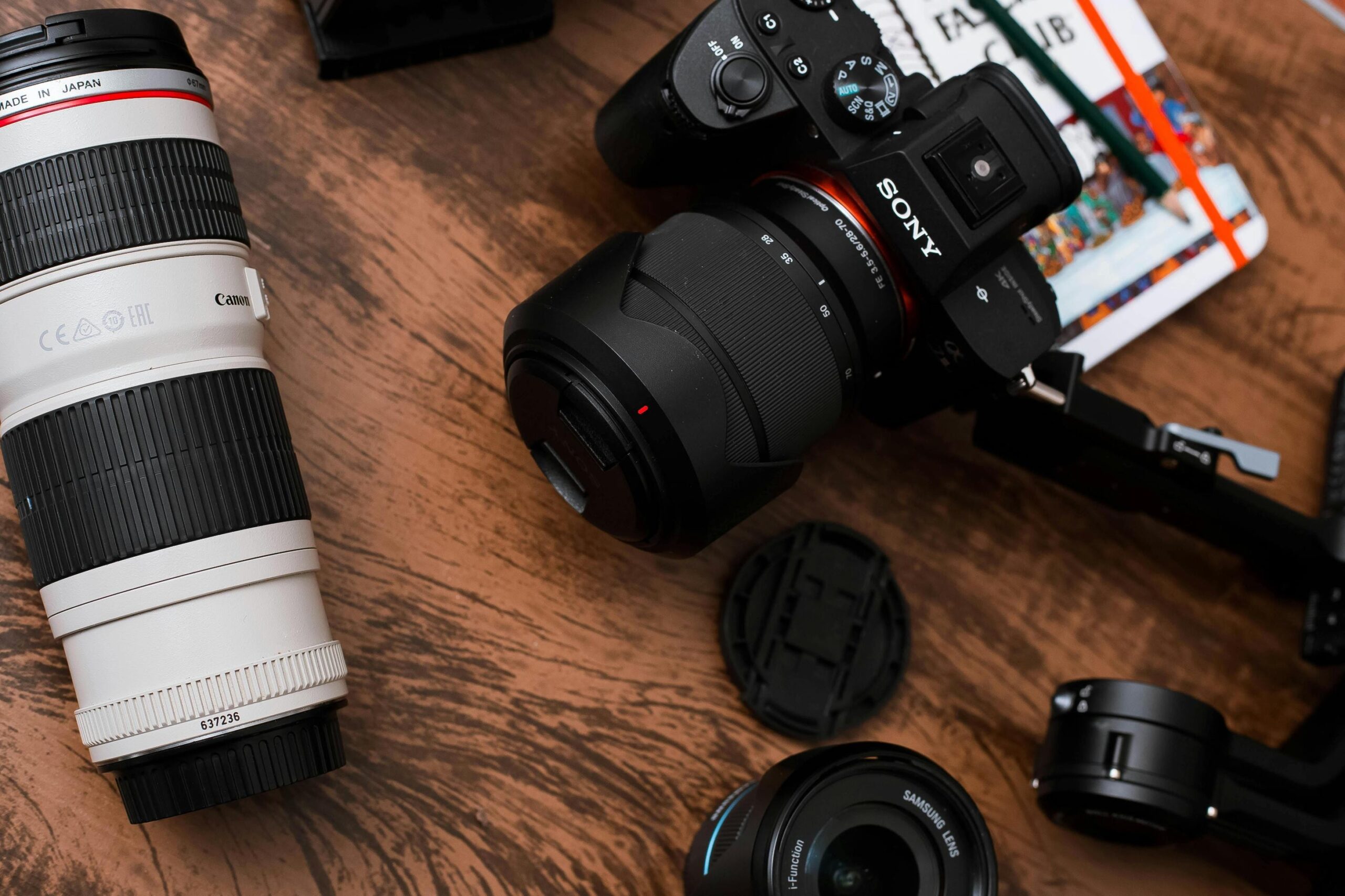  A Sony mirrorless camera equipped with a 28-70mm lens sits on a wooden surface alongside a prominent white Canon telephoto lens. The arrangement includes various accessories such as a Samsung lens, a lens cap, and a notebook with pencils, creating a professional overhead flat-lay.