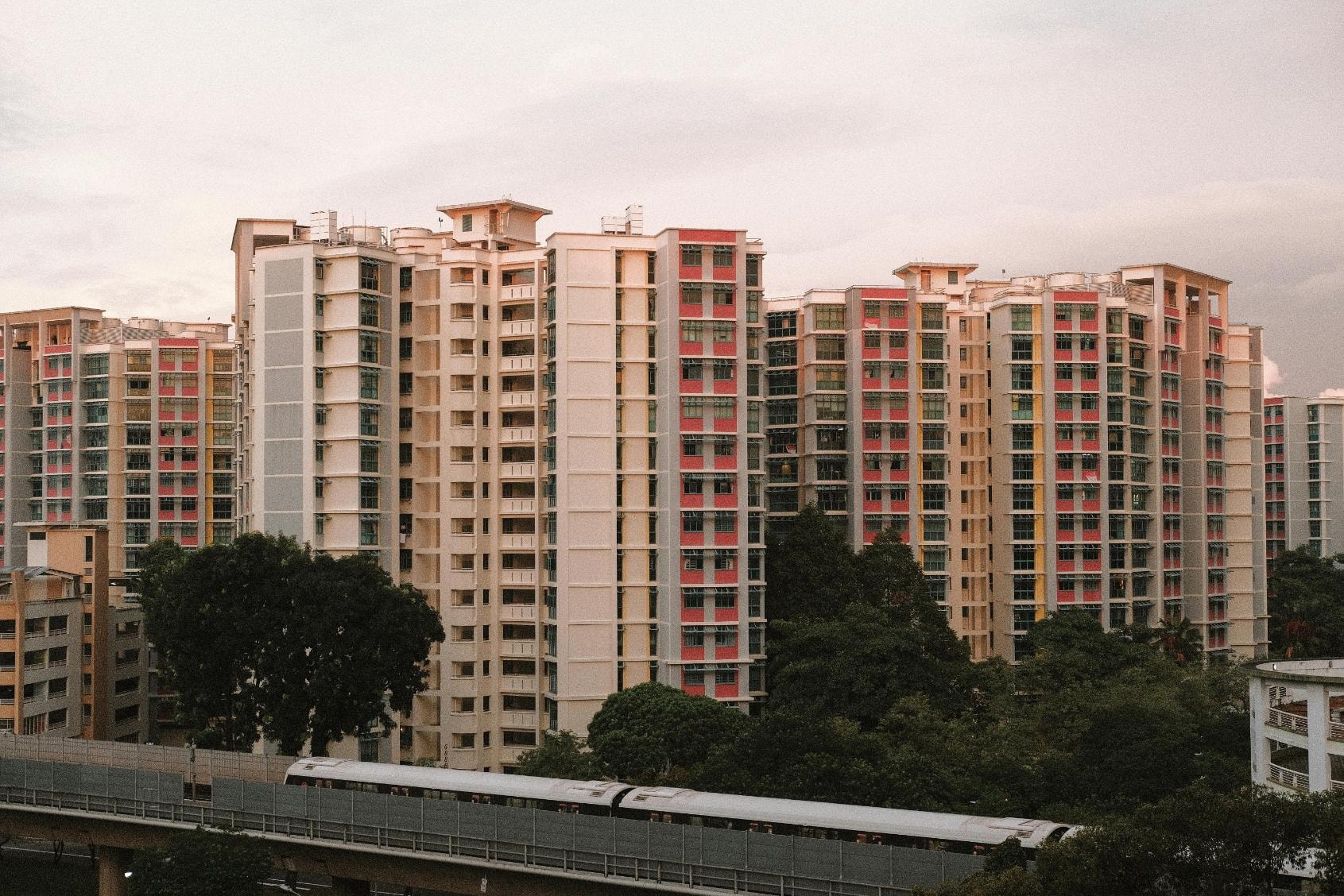 A dense cluster of beige high-rise apartment buildings featuring colorful red and yellow accents dominates the skyline against a soft, overcast sky. In the foreground, lush green trees separate the residential blocks from an elevated railway track where a train is passing.