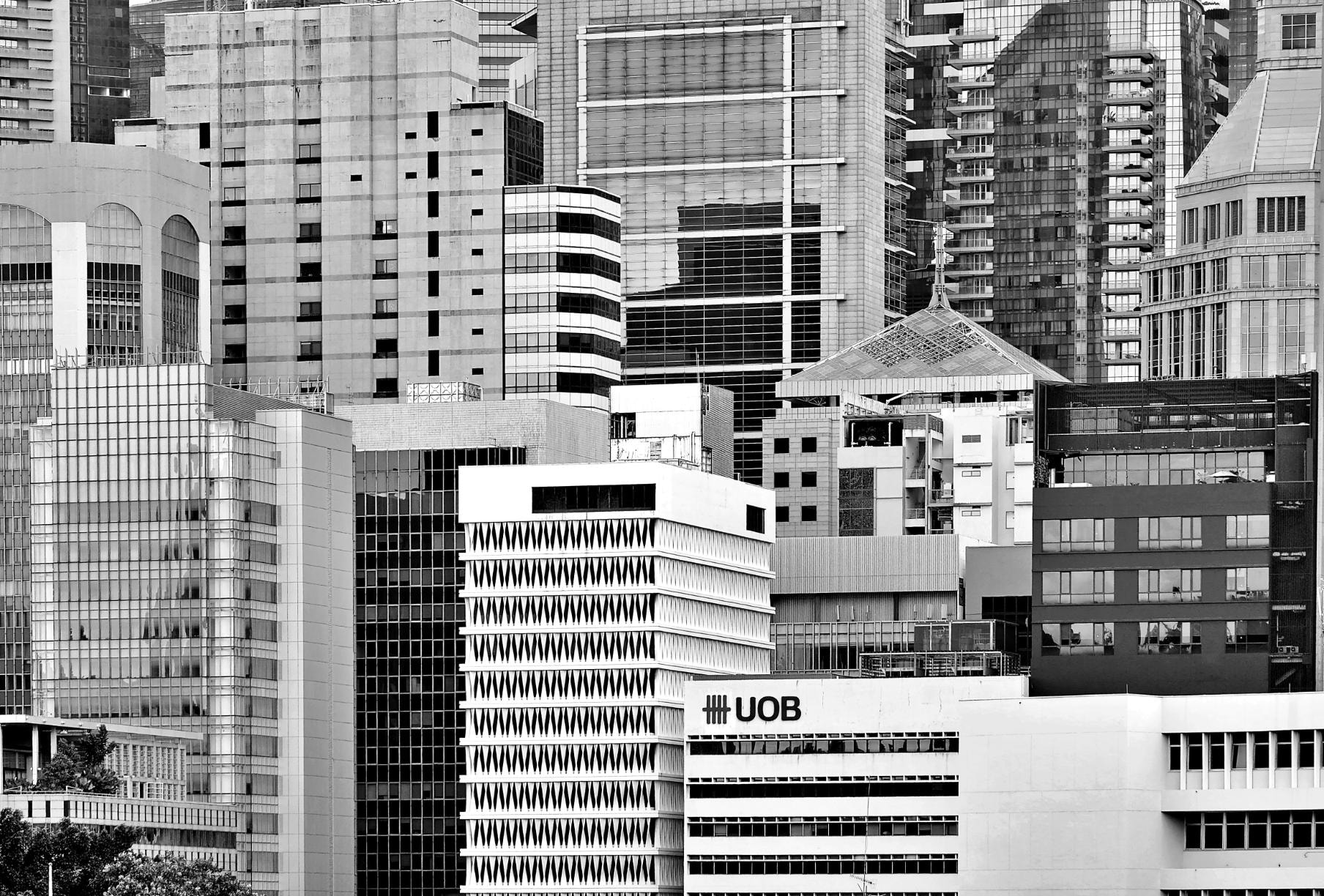 This monochromatic photograph presents a tightly compressed view of a city skyline, stacking layers of skyscrapers to create a dense wall of architectural patterns. Various textures from glass facades and concrete grids interact within the frame, which features a building marked with the UOB logo near the bottom.