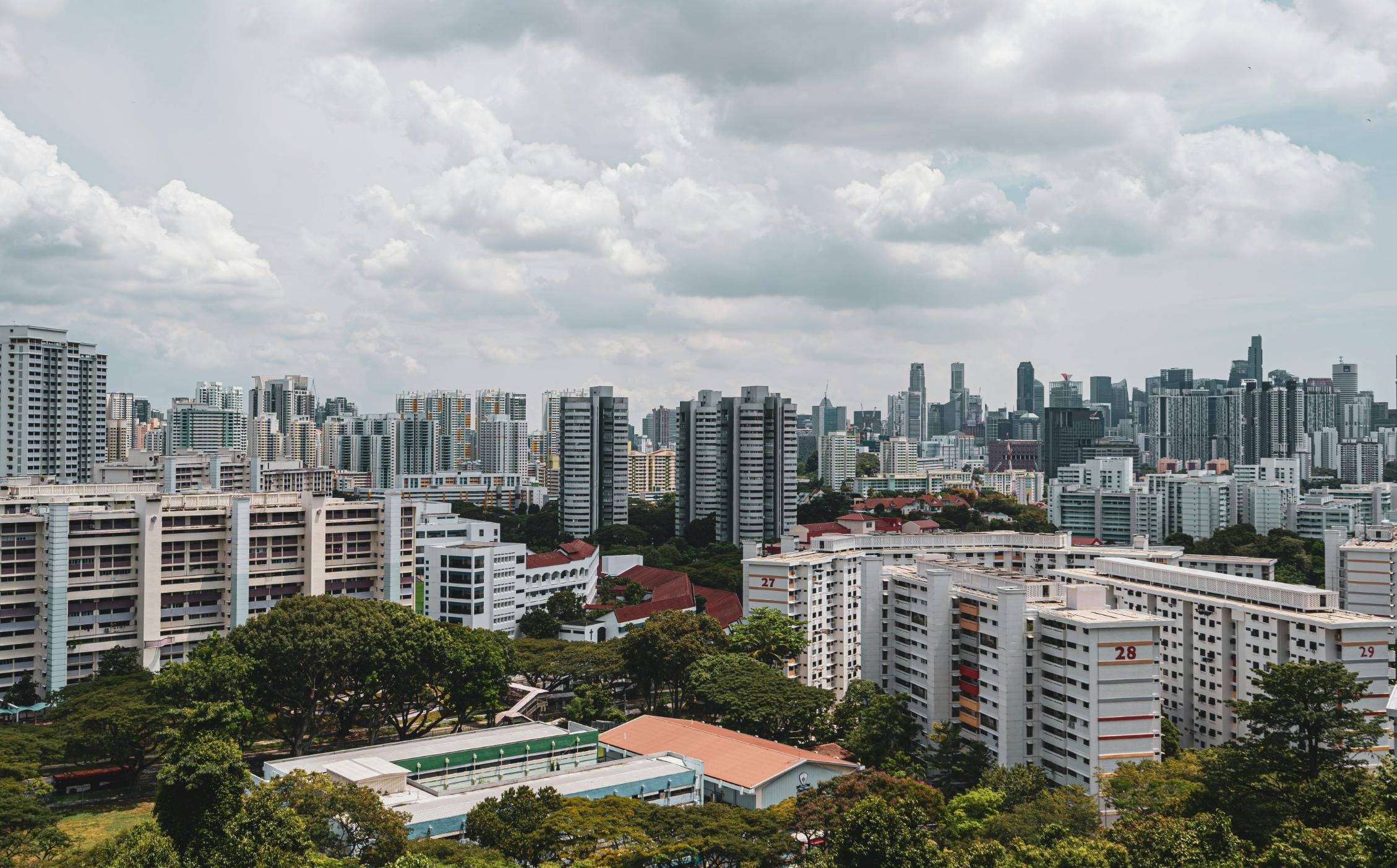 A sweeping urban view features clusters of white residential apartment blocks with red accents nestled among lush greenery in the mid-ground. In the distance, a dense skyline of modern skyscrapers and glass towers stretches across the horizon under a bright, cloudy sky.