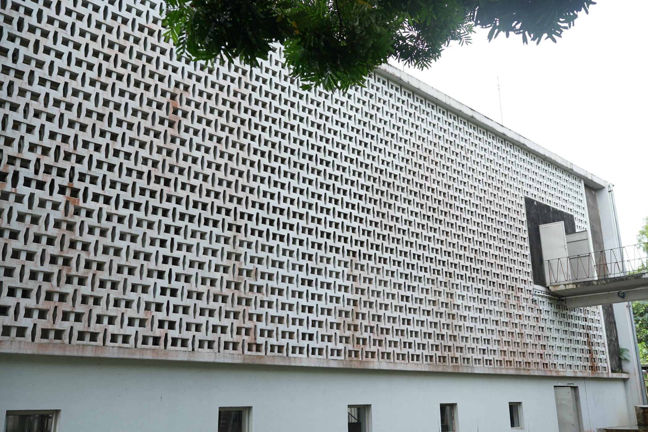 This low-angle photograph captures a building facade defined by a massive upper wall constructed from a repetitive grid of white, geometric breeze blocks. Overhanging green foliage frames the top left corner, contrasting with the structured concrete, while a plain lower wall with small windows and a metal-railed walkway anchors the structure.