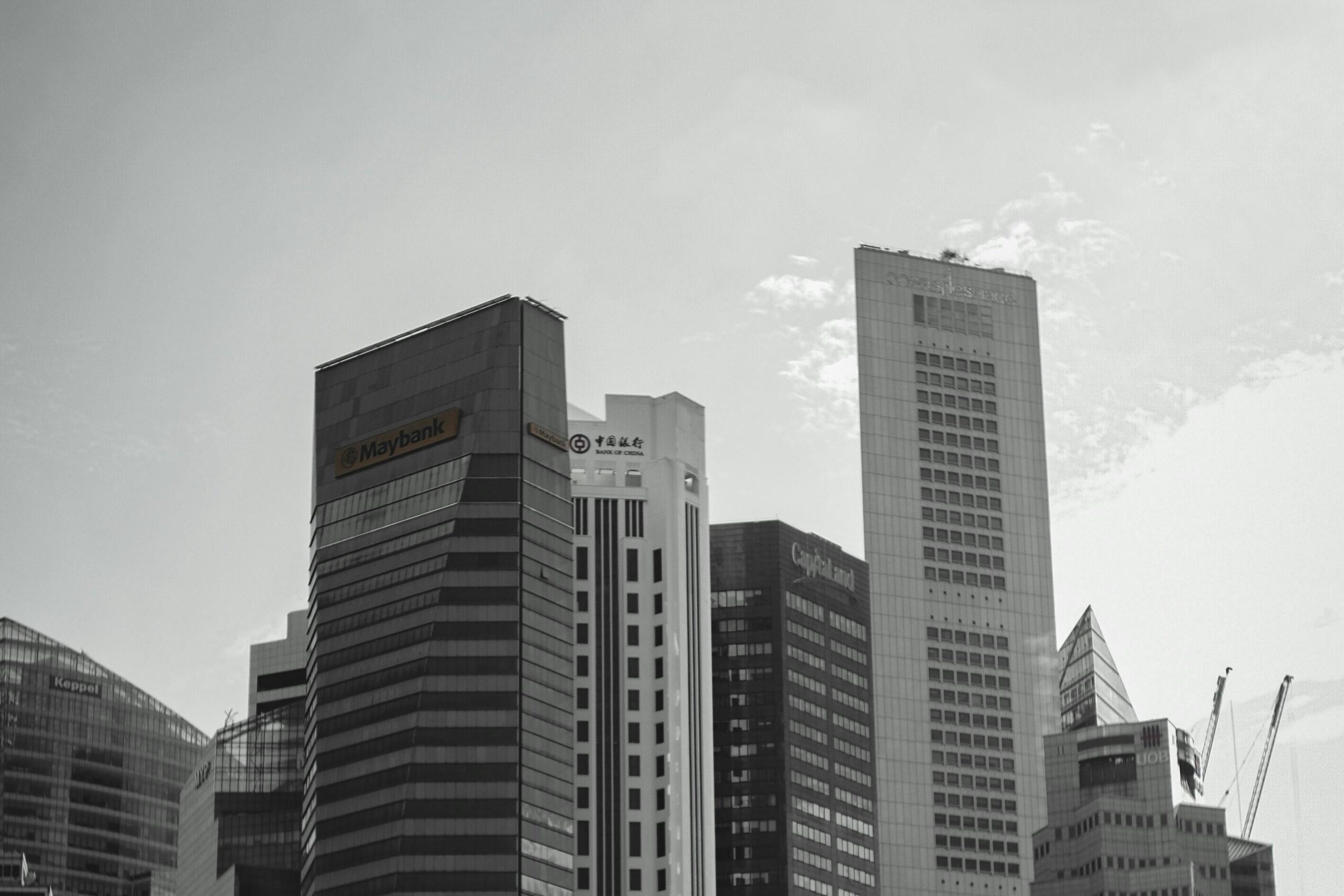 This monochromatic image showcases the skyline of Singapore's central business district, featuring prominent skyscrapers bearing the Maybank, Bank of China, and Capitaland logos. The composition captures the dense clustering of these architectural giants against a bright, cloudy sky, with construction cranes visible on the far right.