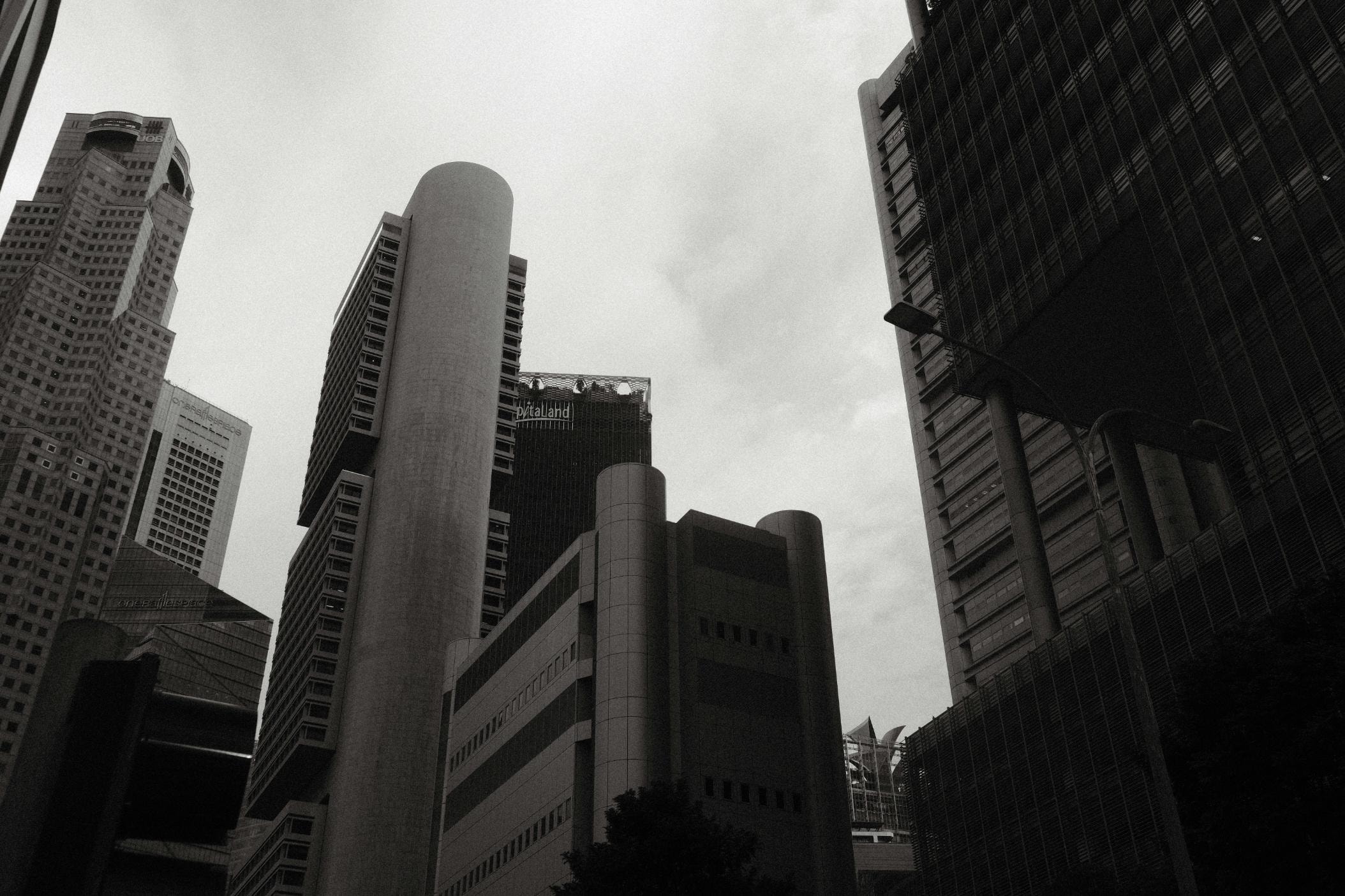 This monochromatic photograph captures a dramatic low-angle view of a dense cluster of skyscrapers, showcasing a mix of curved and geometric architectural styles. The grainy texture and high contrast enhance the imposing verticality of the buildings, creating a moody and atmospheric urban scene against an overcast sky.