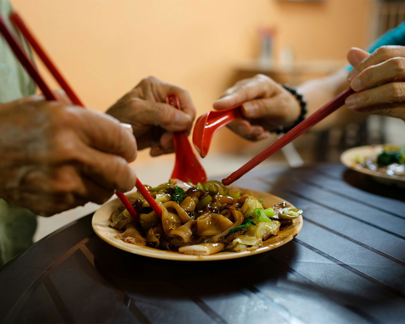 Two hands holding chopsticks lift strands of fried kueh tiew from a shared plate, showing the moment of connection and casual sharing over a simple local meal.