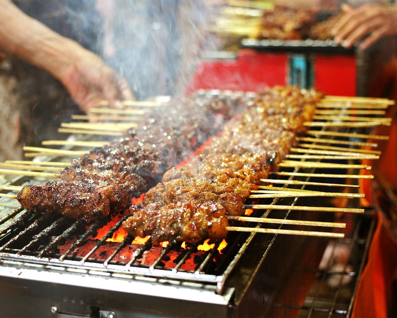 Close-up view of satay skewers sizzling over open flames, with smoke rising and meat glistening, highlighting the texture and aroma of freshly grilled street food.