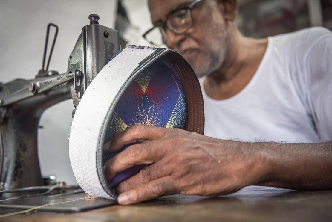 An elderly artisan wearing glasses meticulously guides a patterned songkok through a vintage sewing machine. His weathered hands focus on the intricate stitching of the traditional headwear, which features a vibrant blue and red interior design.