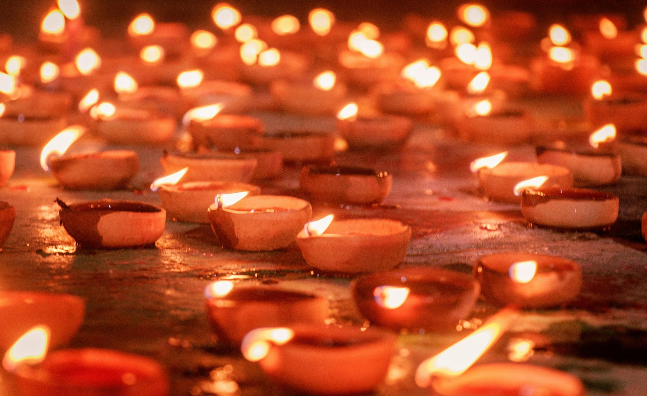 Countless small clay oil lamps, known as diyas, are arranged across a ground surface, casting a warm and flickering orange glow. The shallow depth of field creates a soft bokeh effect in the background, emphasizing the repetitive, rhythmic beauty of the traditional lights.