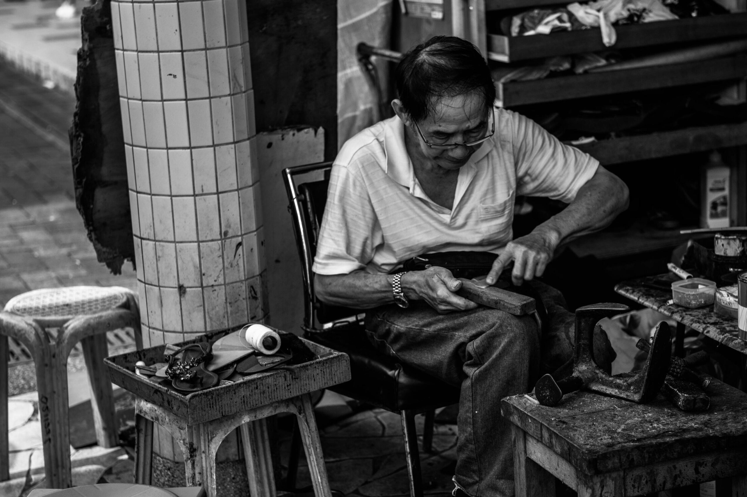 A cobbler is captured in a black-and-white photograph, focused on repairing footwear at his outdoor workstation. He sits amidst his tools and materials, including a shoe last and a tray of supplies, while working on a piece of leather.