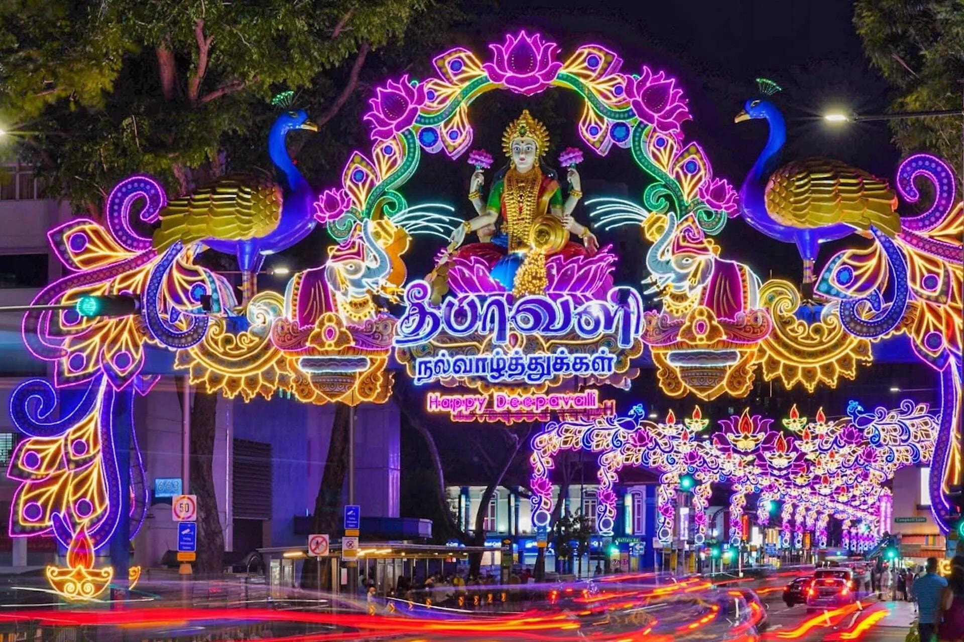 A dazzling light display for Deepavali spans a city street, featuring a central figure of a goddess flanked by majestic peacocks and elephants. Below the ornate, neon-lit arch, blurred light trails from moving traffic create a vibrant, high-energy atmosphere through the heart of the celebration.