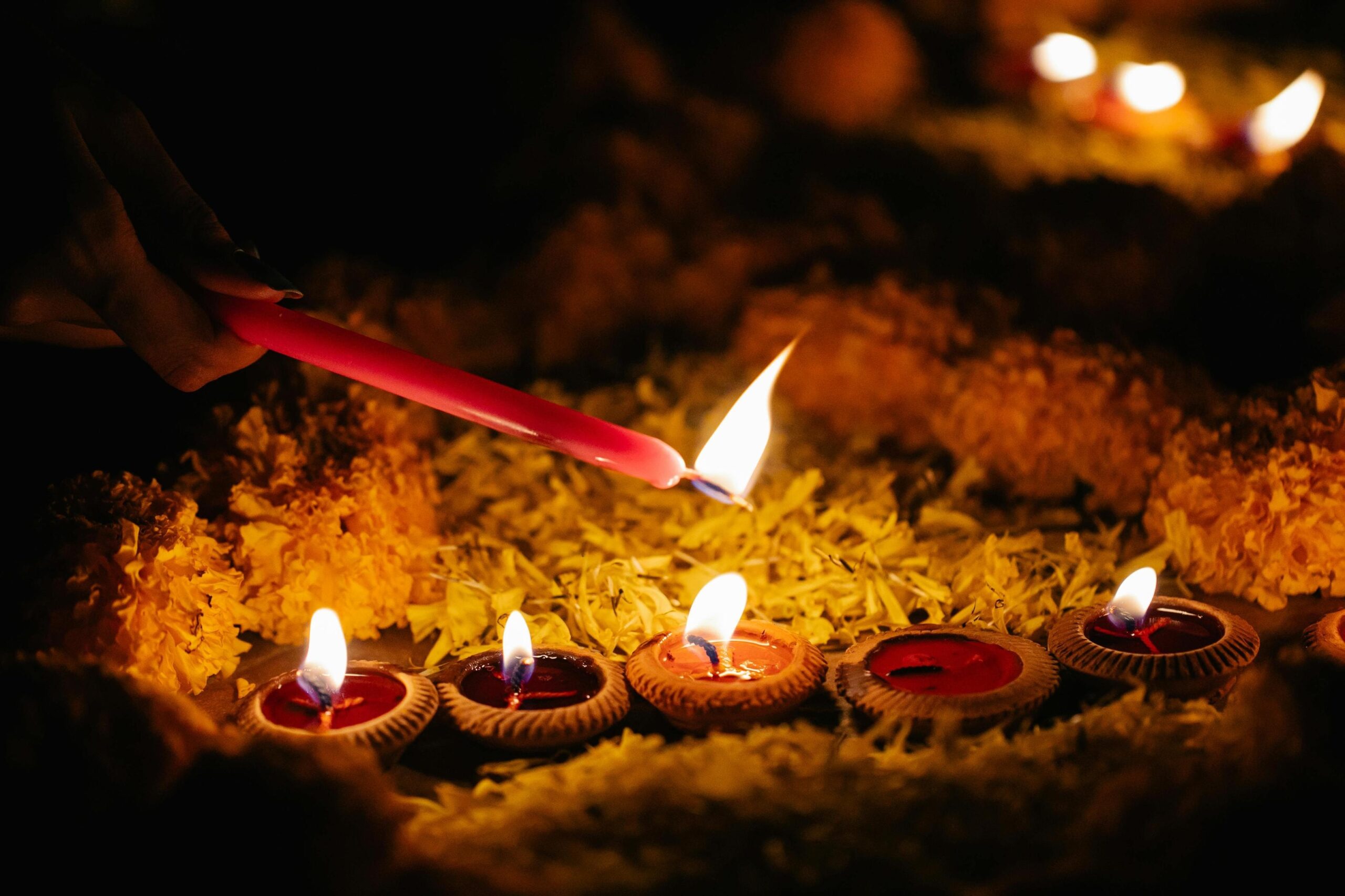 A person's hand uses a slender pink candle to light a series of small, traditional clay lamps nestled among bright yellow flowers. The warm glow of the flickering flames illuminates the surrounding marigolds, creating a peaceful and ceremonial atmosphere against the dark background.