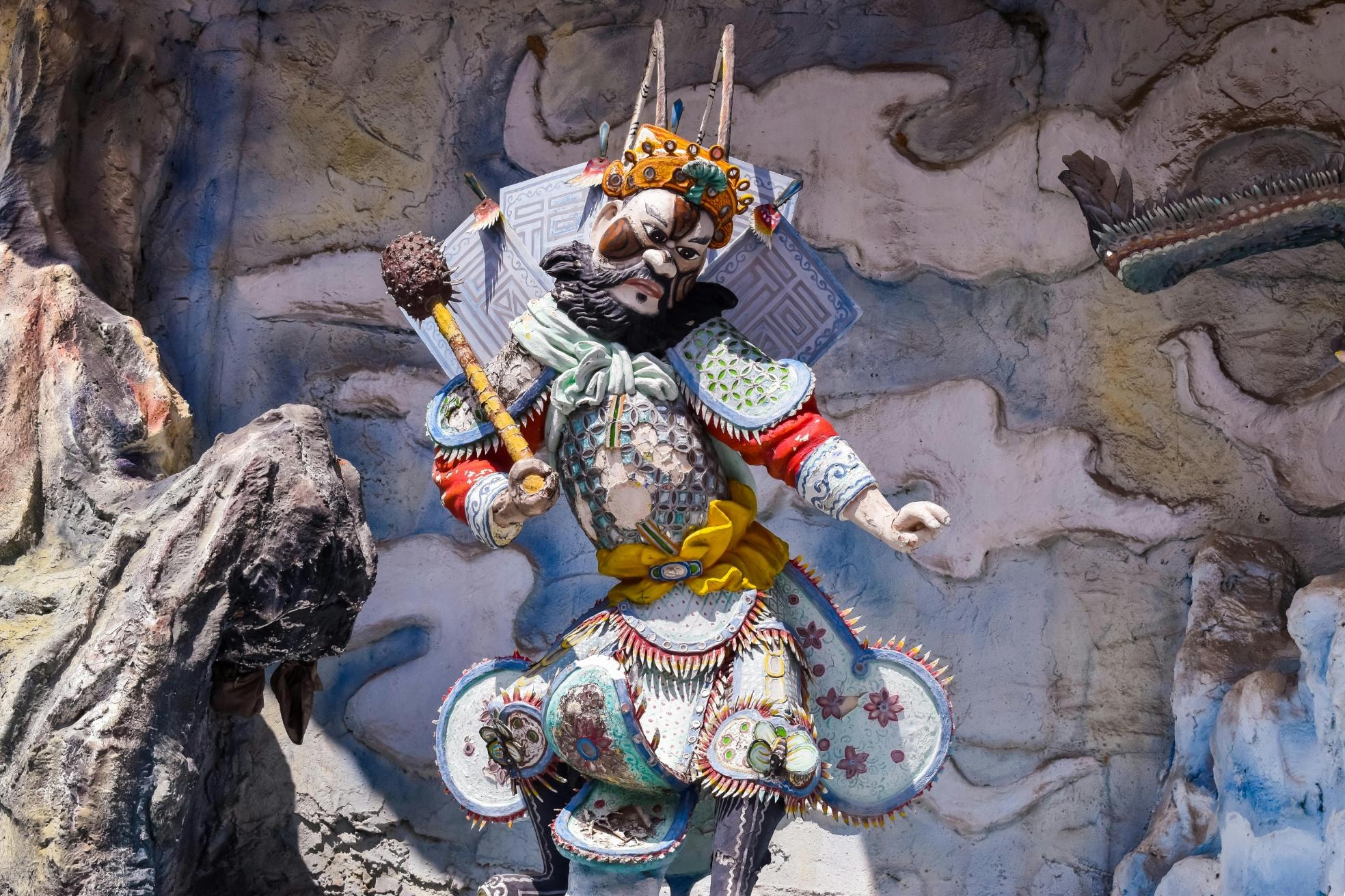 An ornate, bearded warrior statue stands holding a mace against a textured wall featuring sculpted clouds and rock formations. The figure is dressed in vibrant, traditional armor detailed with intricate patterns, a yellow sash, and a spiked crown.