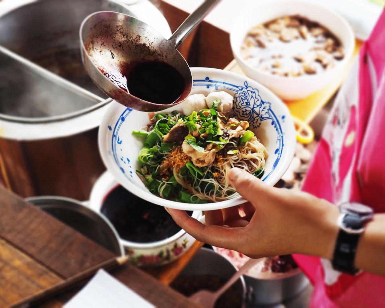 A hand pours hot soup into a bowl of mee, with steam rising gently, capturing the final step before serving a comforting bowl of noodles.