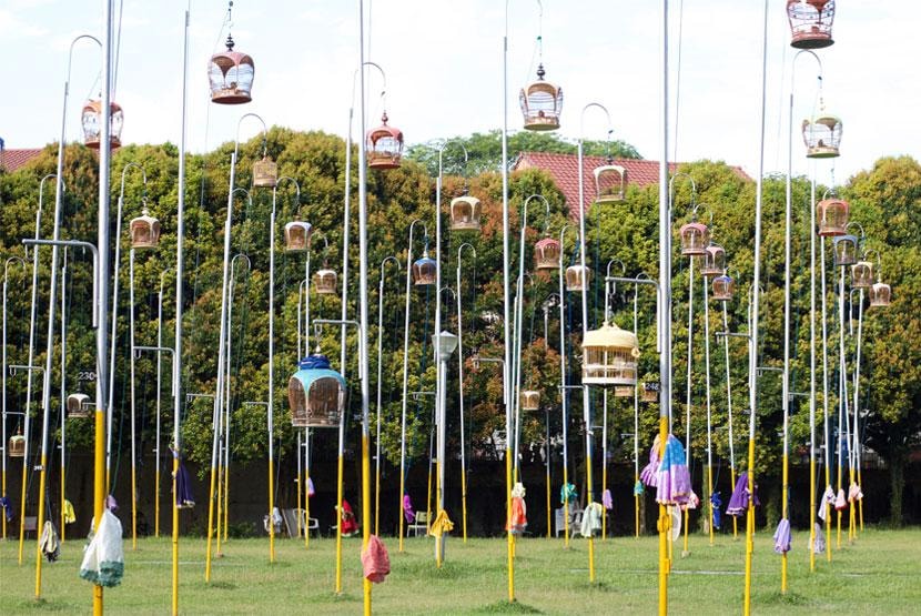 Numerous decorative birdcages are hoisted high on slender metal poles across a grassy field, often part of traditional birdsong competitions. Colorful cloths hang from the base of several poles against a backdrop of dense green trees and a red-tiled roof.