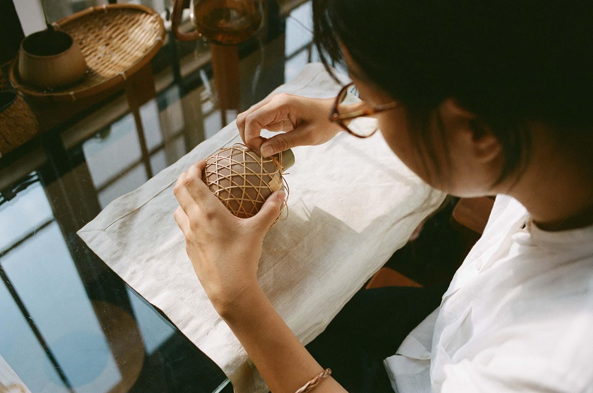 A young woman wearing glasses is shown from an over-the-shoulder perspective as she carefully weaves a delicate bamboo lattice around a small ceramic vessel. Sitting at a glass table bathed in natural light, she demonstrates the precision and focus required for this traditional craft.