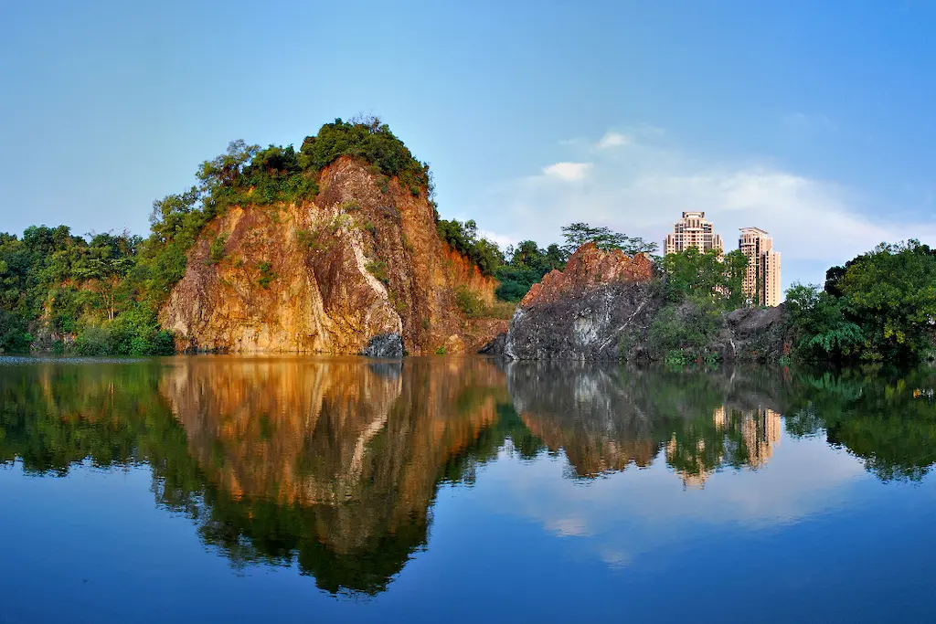 A rocky cliff covered in green vegetation rises from the edge of a calm lake, creating a vibrant reflection on the water's surface. In the background, modern high-rise buildings peek through the trees under a clear blue sky, blending nature with urban development.