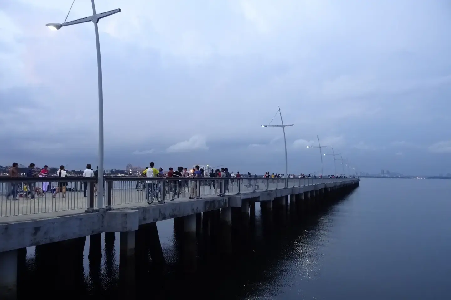 A crowd of people gathers along a long, concrete pier stretching out into a calm body of water under a twilight sky. Modern, minimalist street lamps line the walkway, casting a soft glow as the horizon fades into deep blue and grey tones.