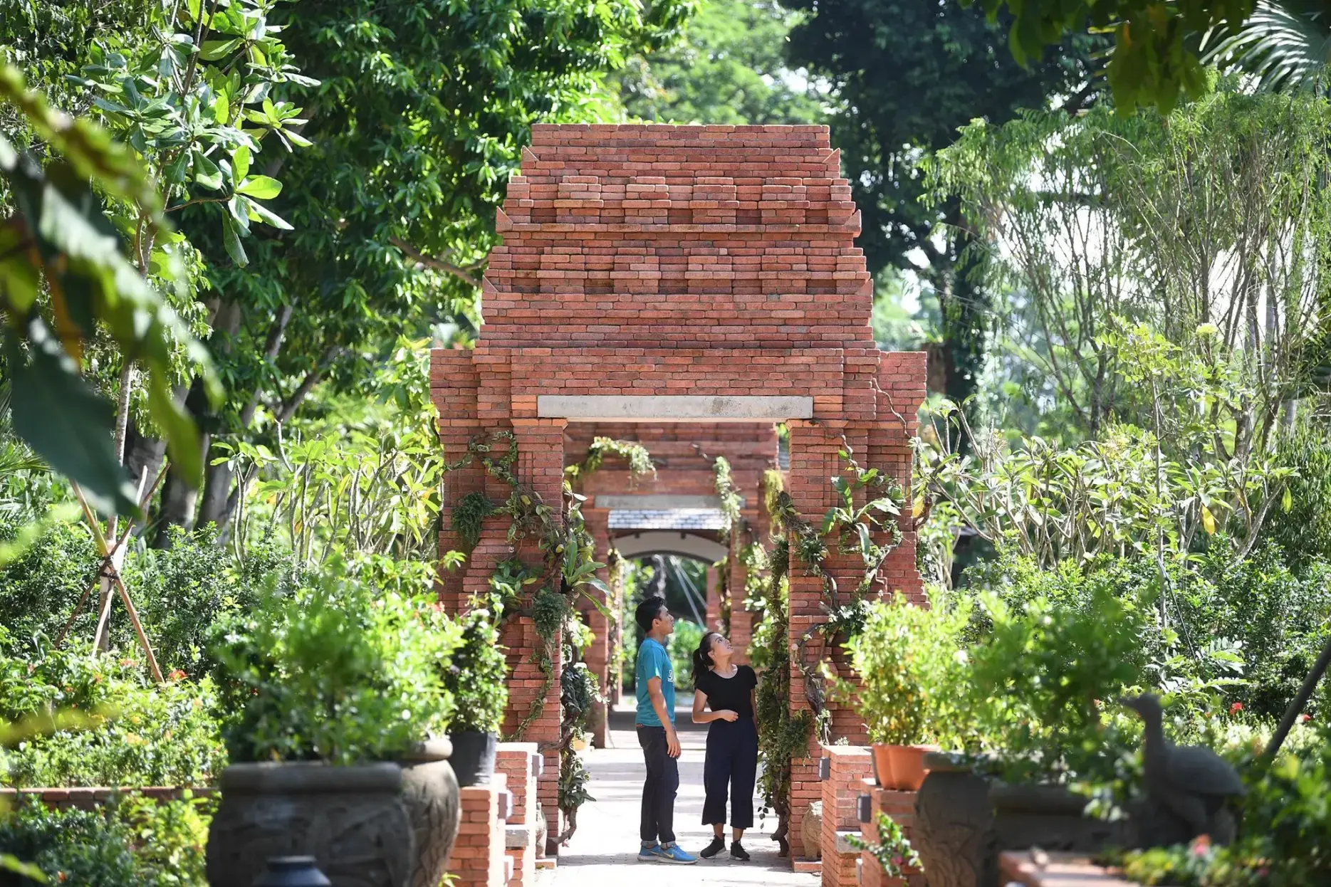 A man and a woman stand within a lush garden, looking up at a series of towering, tiered red brick archways. Sunlight filters through the surrounding green trees and potted plants, illuminating the stone path that leads through the architectural structures.