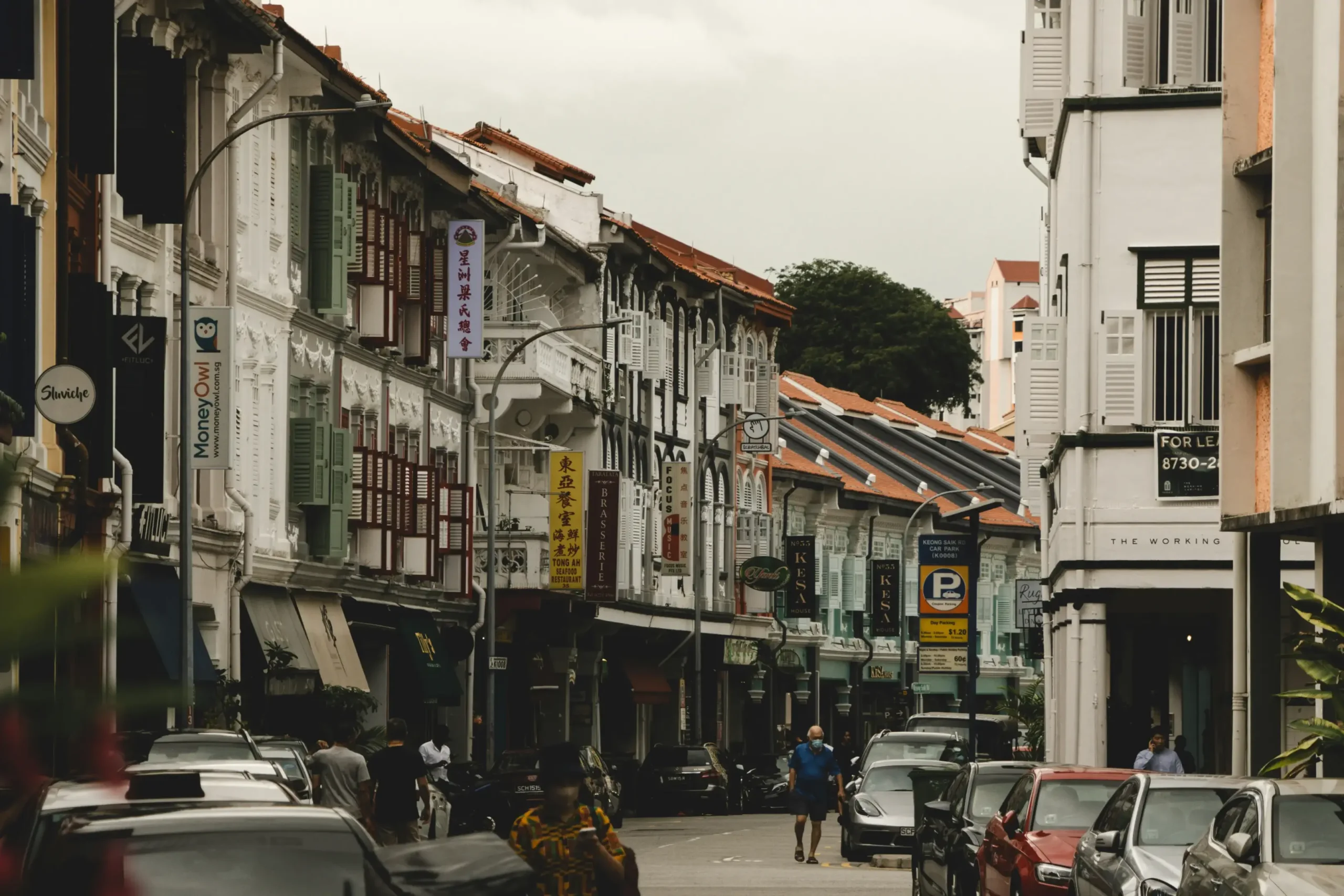 This image captures a narrow, busy street lined with traditional colorful shophouses featuring ornate shutters and vintage architectural details. Numerous business signs in various languages hang above the parked cars and pedestrians moving along the road.