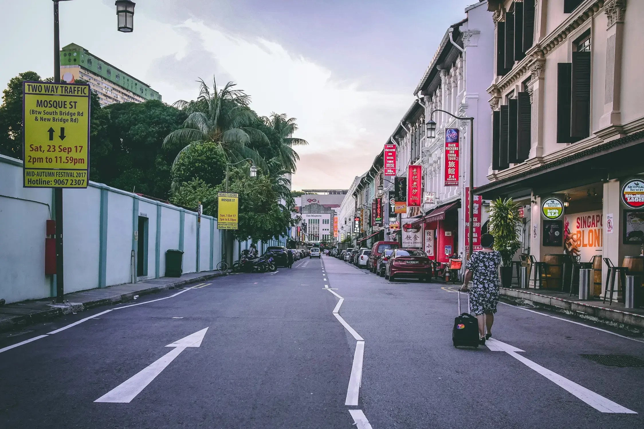 A person pulls a suitcase down the middle of Mosque Street in Singapore, which is lined with colorful shopfronts and parked cars. A bright yellow traffic sign on the left provides details for a festival, set against a backdrop of lush trees and a pale evening sky.