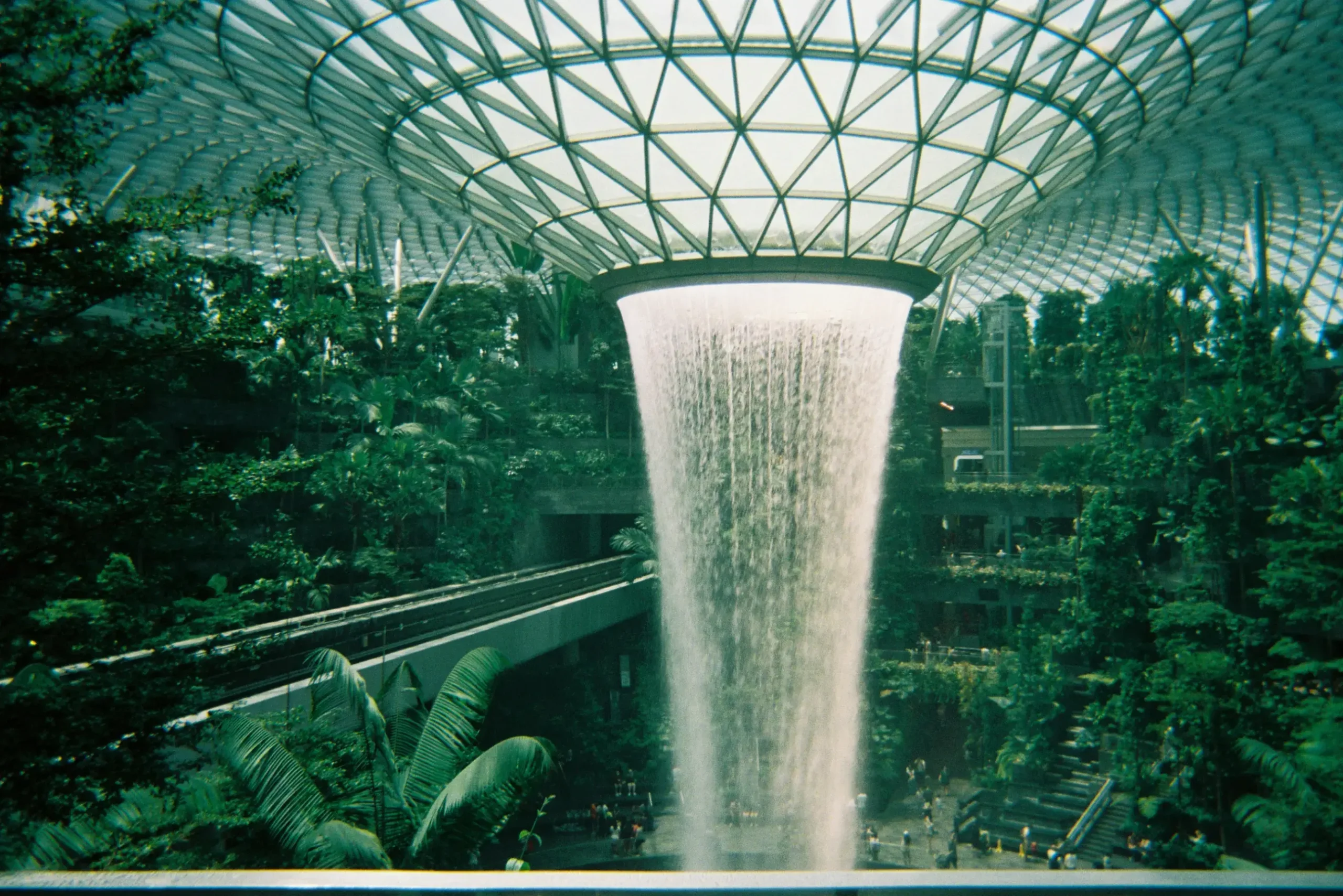 A massive indoor waterfall plunges from a geometric glass dome into a lush, multi-level forest filled with tropical greenery. The architecture seamlessly blends nature with modern design, featuring a transit track that winds through the dense foliage.