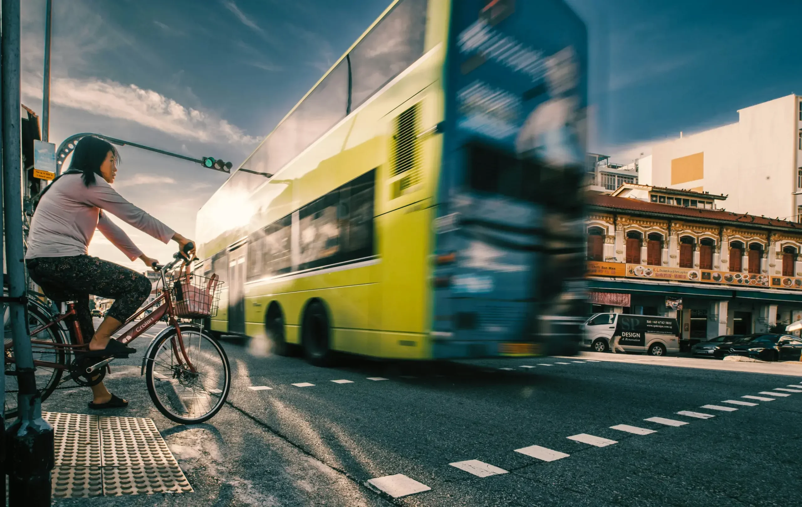 A woman on a bicycle waits at a street corner as a lime-green double-decker bus speeds past her in a blur of motion. The urban scene is captured during the golden hour, highlighting traditional shophouses in the background under a clear, sunlit sky.