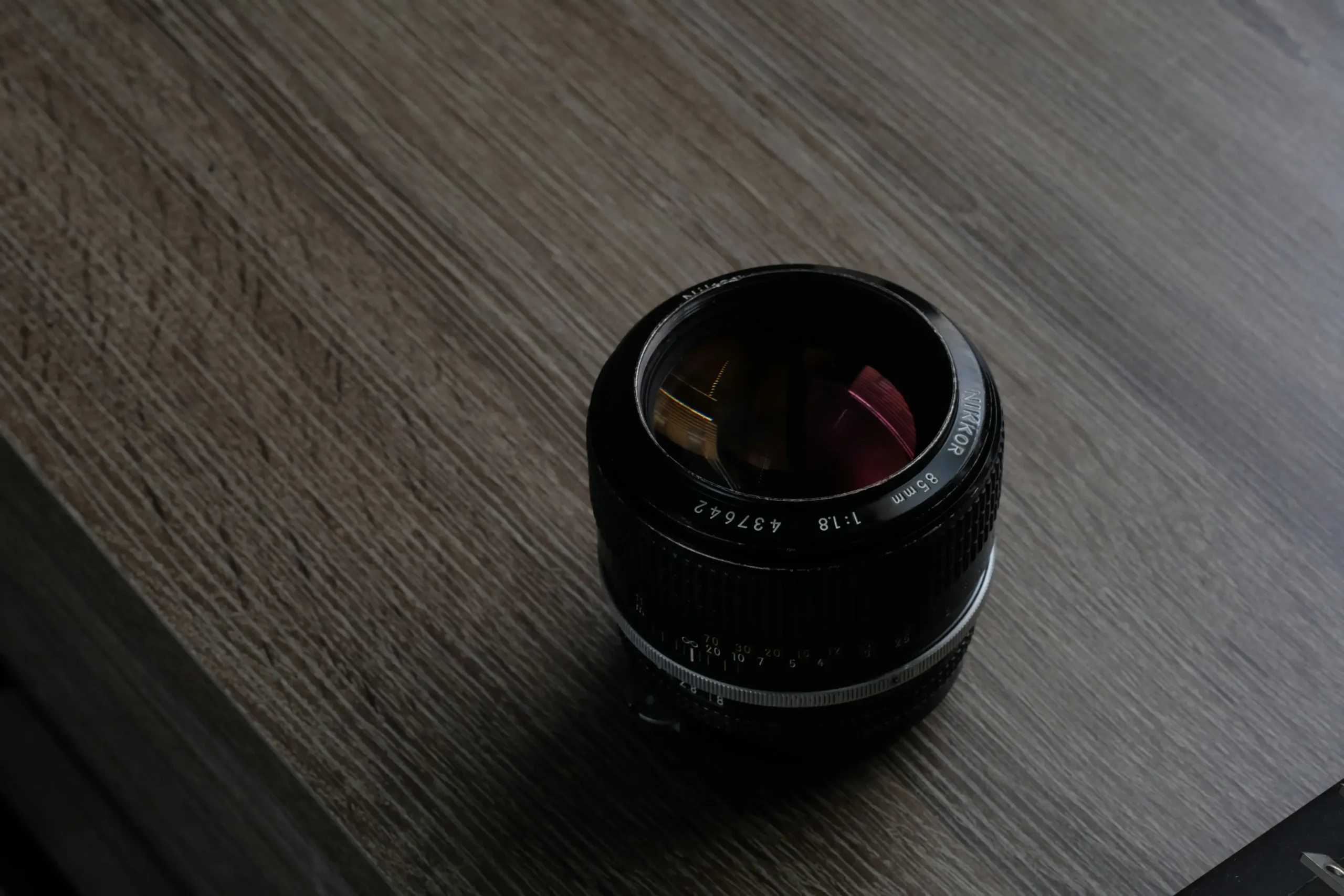 A vintage Nikkor 85mm f/1.8 camera lens sits centered on a grey, wood-grained surface. The shot features a high-angle perspective, highlighting the lens's glass reflections and metallic focus rings.