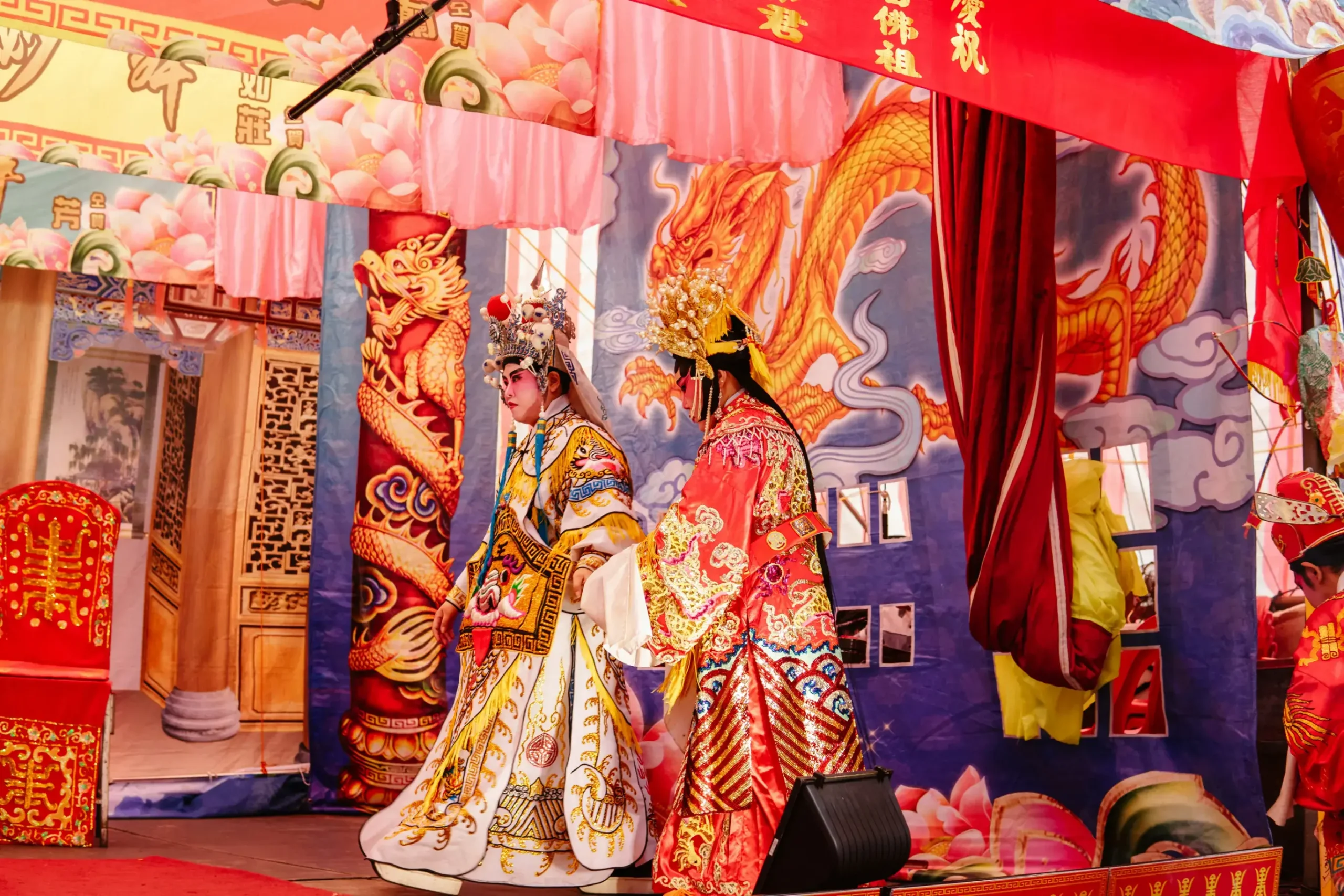 Two performers dressed in elaborate, colorful traditional Chinese opera costumes stand on a stage decorated with vibrant dragon motifs and banners. The scene captures a moment of dramatic flair, highlighted by the intricate headpieces and the rich, golden embroidery of their attire.