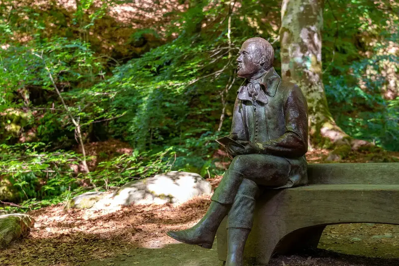 A bronze statue of the Scottish poet Robert Burns sits contemplatively on a wooden bench, holding a small book in his hands. The sculpture is nestled within a lush, sun-dappled forest, surrounded by green foliage and mature trees.